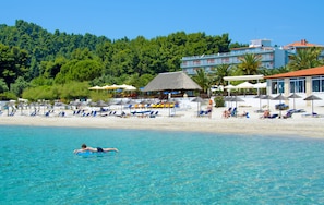 A beach with clear blue water, sun loungers, umbrellas, and a building with a red roof in the background.