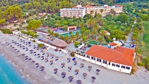 Vista aérea de un hotel costero con varios pisos, una playa con sombrillas y tumbonas, y una piscina rodeada de palmeras.
