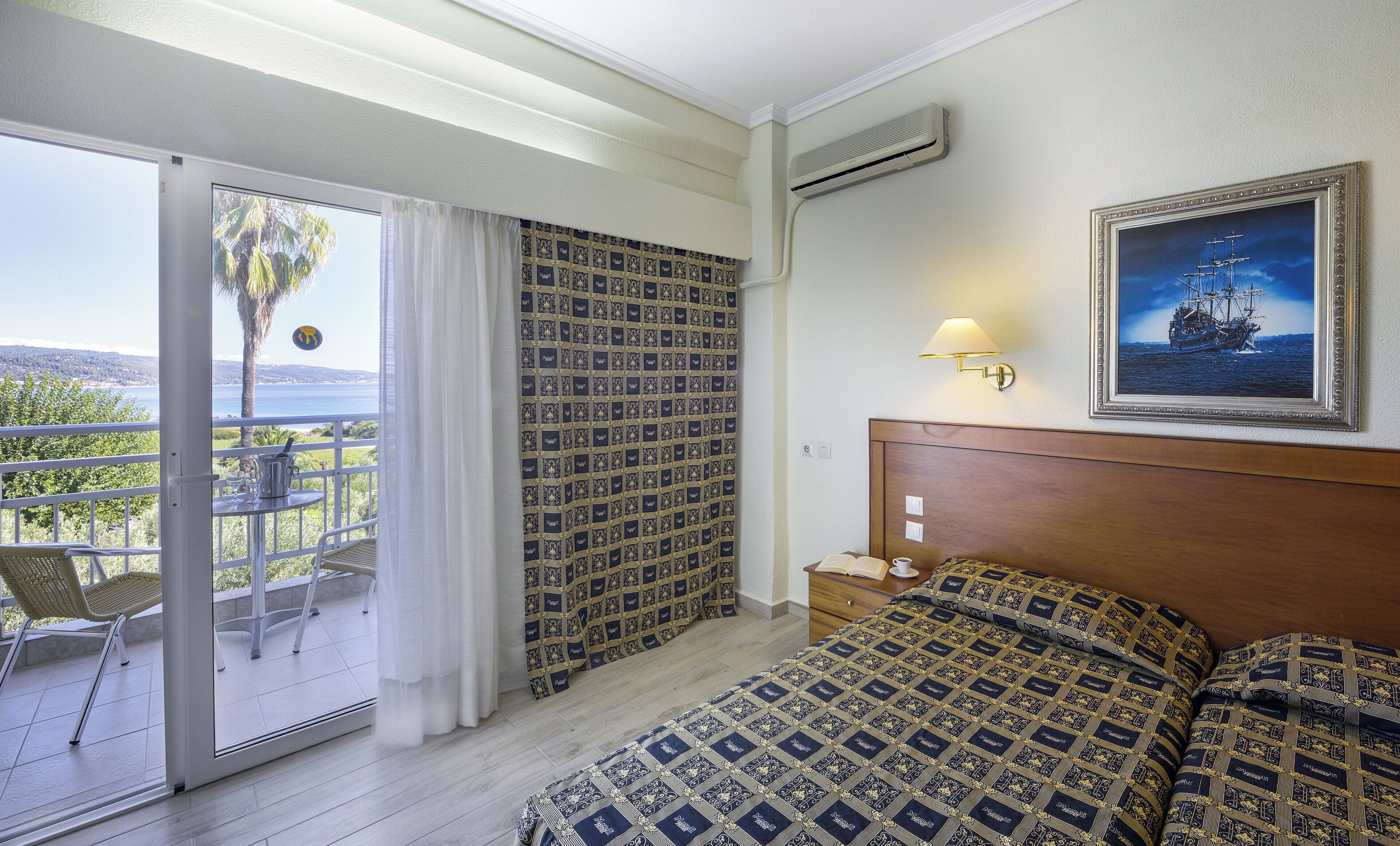 Una habitación de hotel con balcón, cama con ropa de cama estampada, un cuadro en la pared y una mesita de noche.