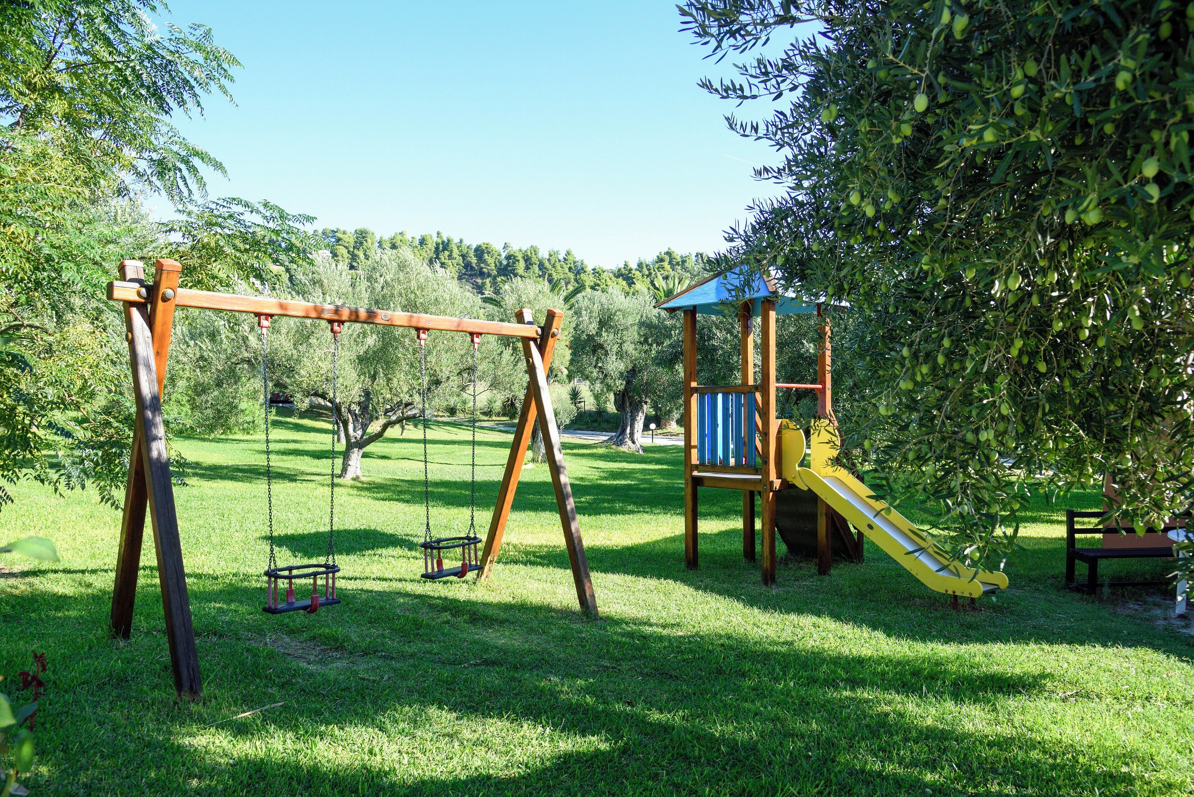 Un parque infantil con columpios y un tobogán, rodeado de árboles y césped.