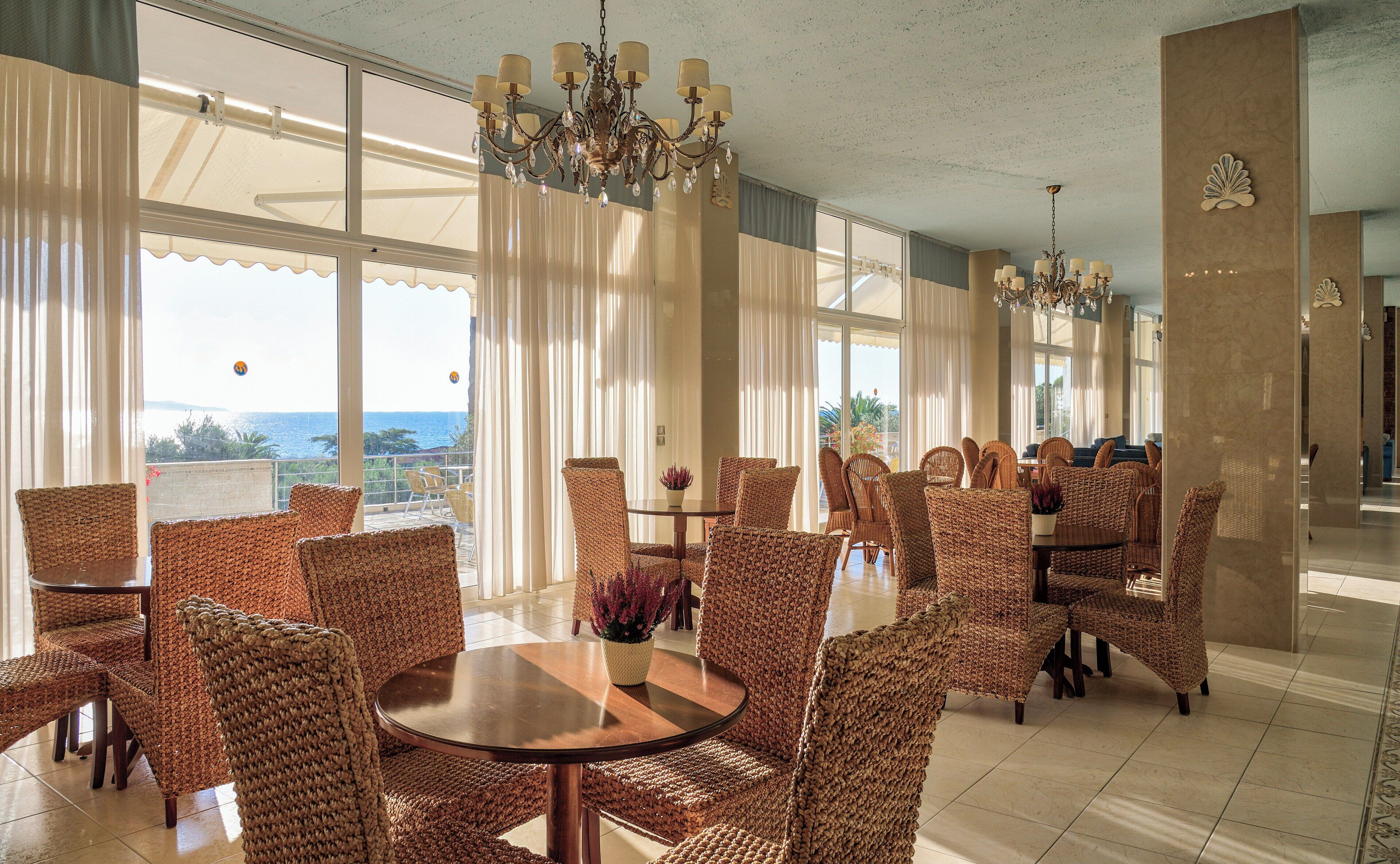 Un comedor amplio con sillas de mimbre y una mesa redonda central, ventanas grandes con vistas al mar y elegantes lámparas de araña.