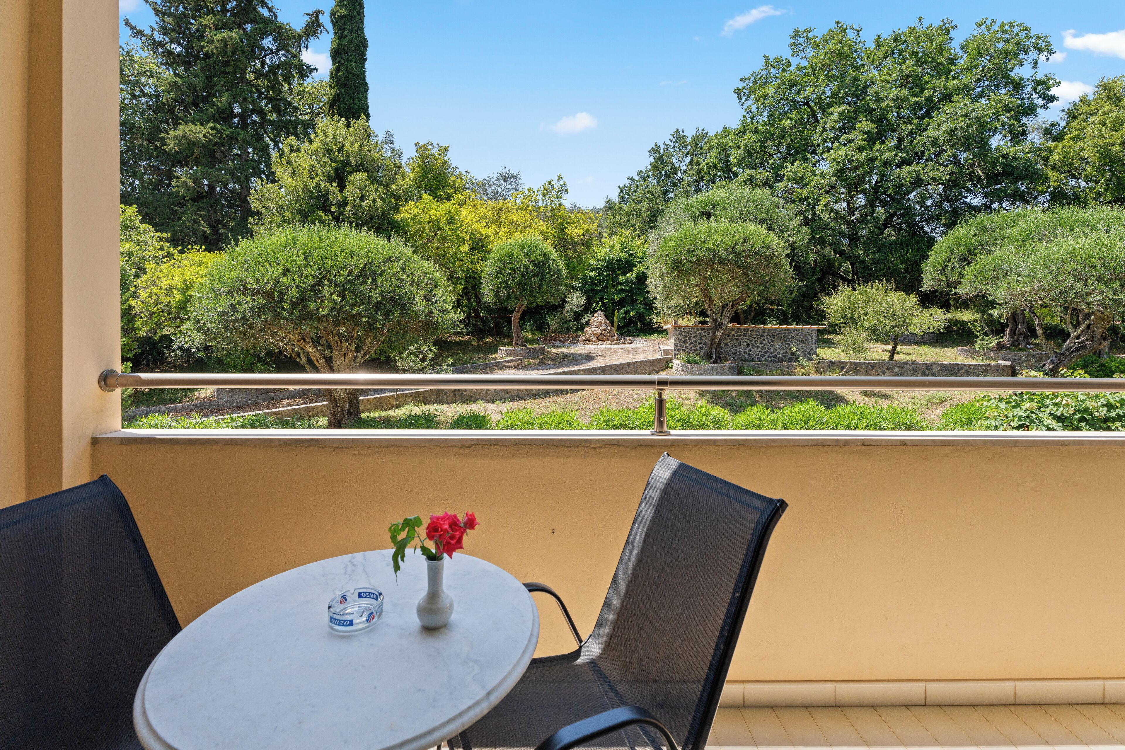 A balcony with a round table and two chairs, overlooking a landscaped garden with various trees and shrubs.