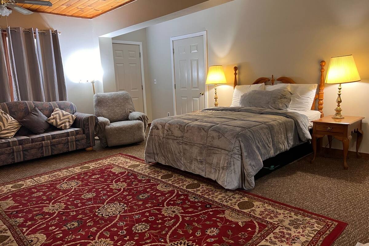 A bedroom with a bed, two bedside tables with lamps, a chair, a sofa, and a red patterned rug.