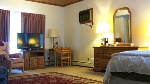 Habitación de hotel con techo de madera, televisor, escritorio con lámpara, una silla, cómoda, cama y alfombra roja con estampado.