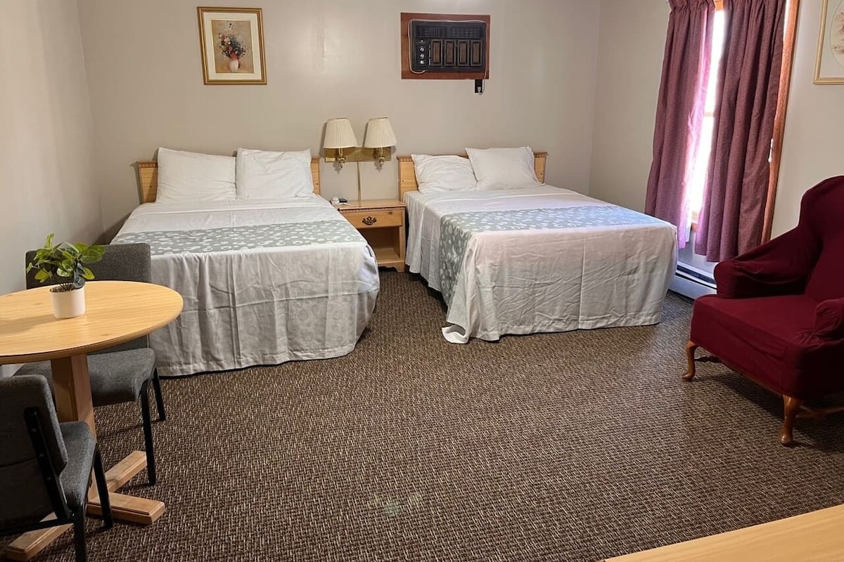 A hotel room with two beds, a dining table, a red armchair, a ceiling fan, and a window with curtains.