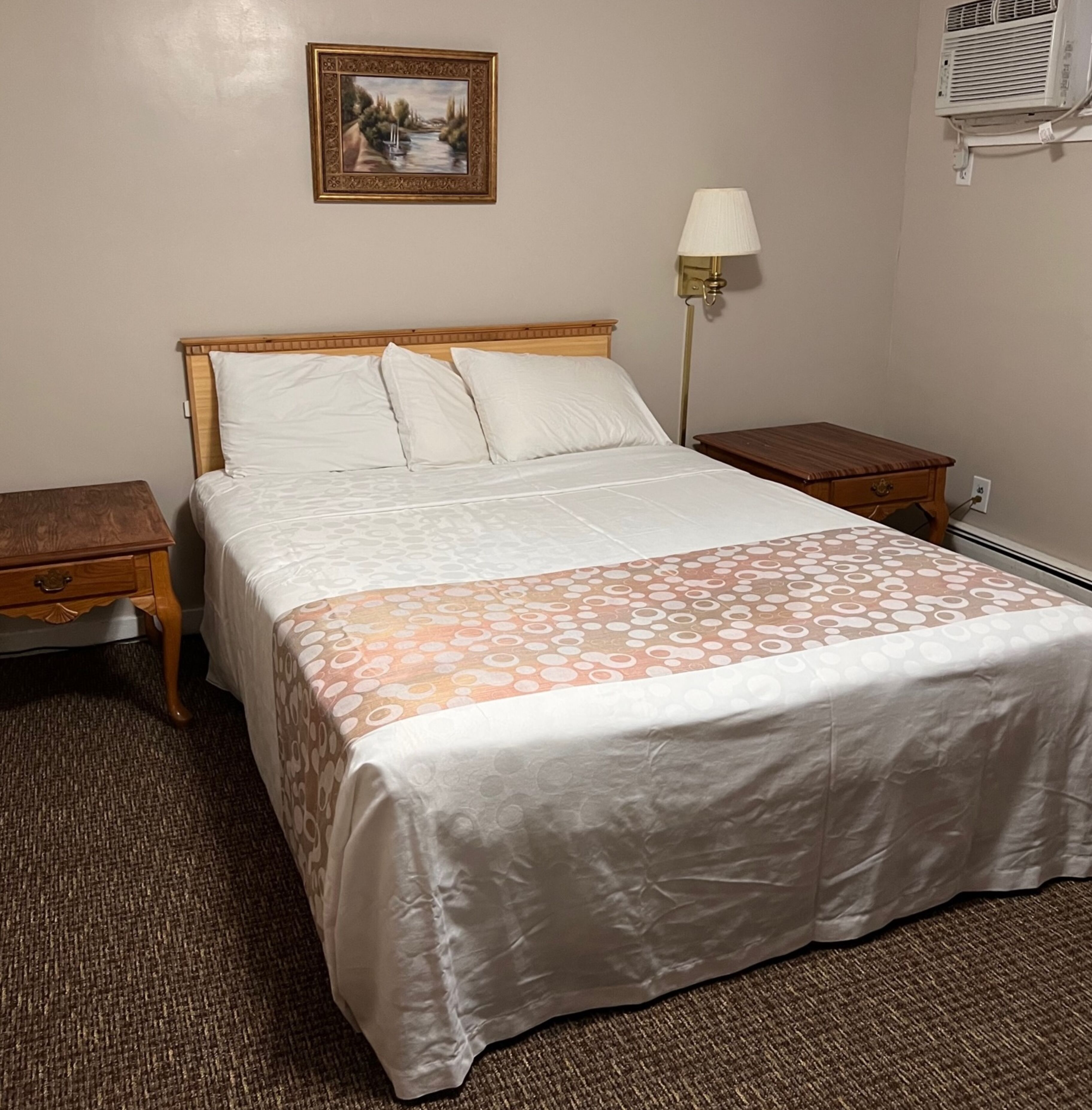 A neatly made bed with a patterned bedspread, a bedside table, a lamp, and a wall-mounted air conditioner.