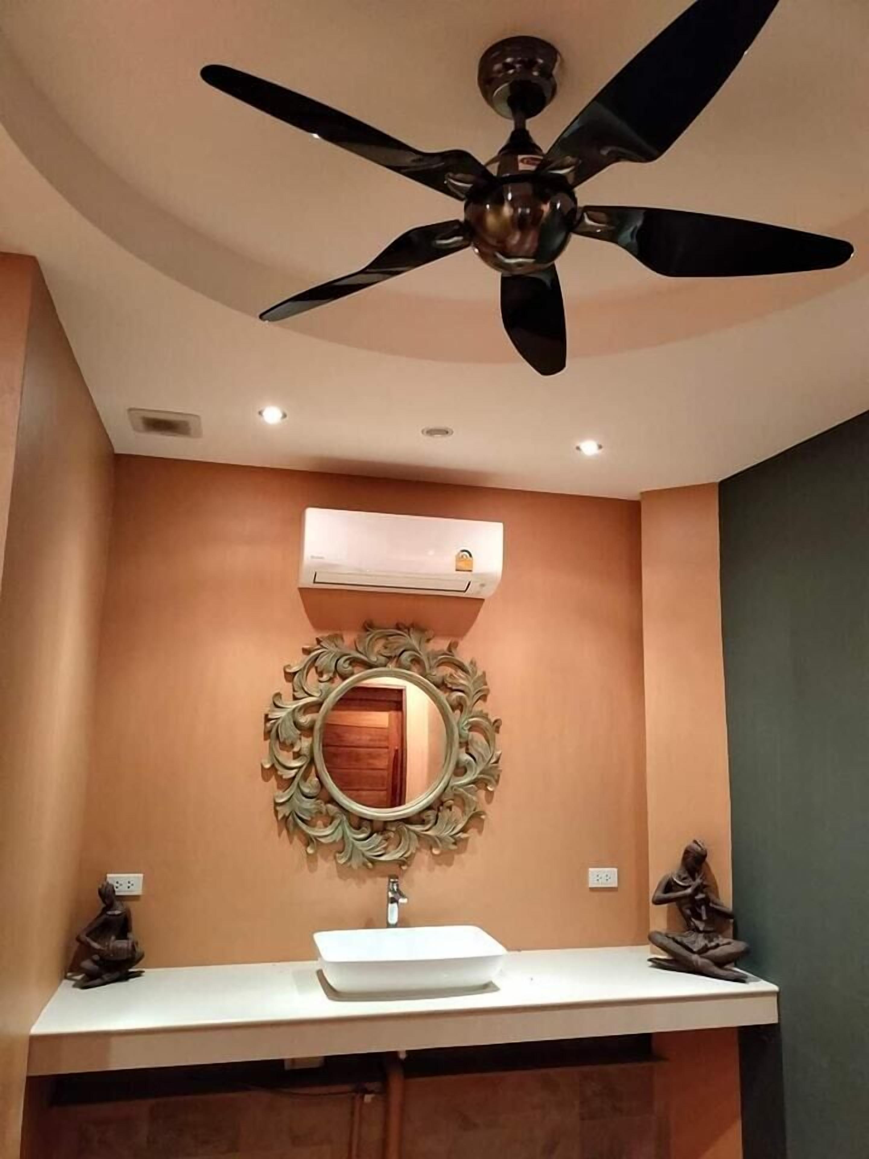 A bathroom with a ceiling fan, a wall-mounted air conditioner, a decorative mirror, a sink, and a statue.
