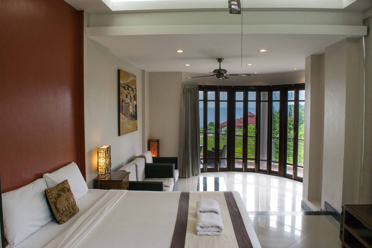 A spacious bedroom with a large bed, a ceiling fan, and a view of the outdoors through a large window.