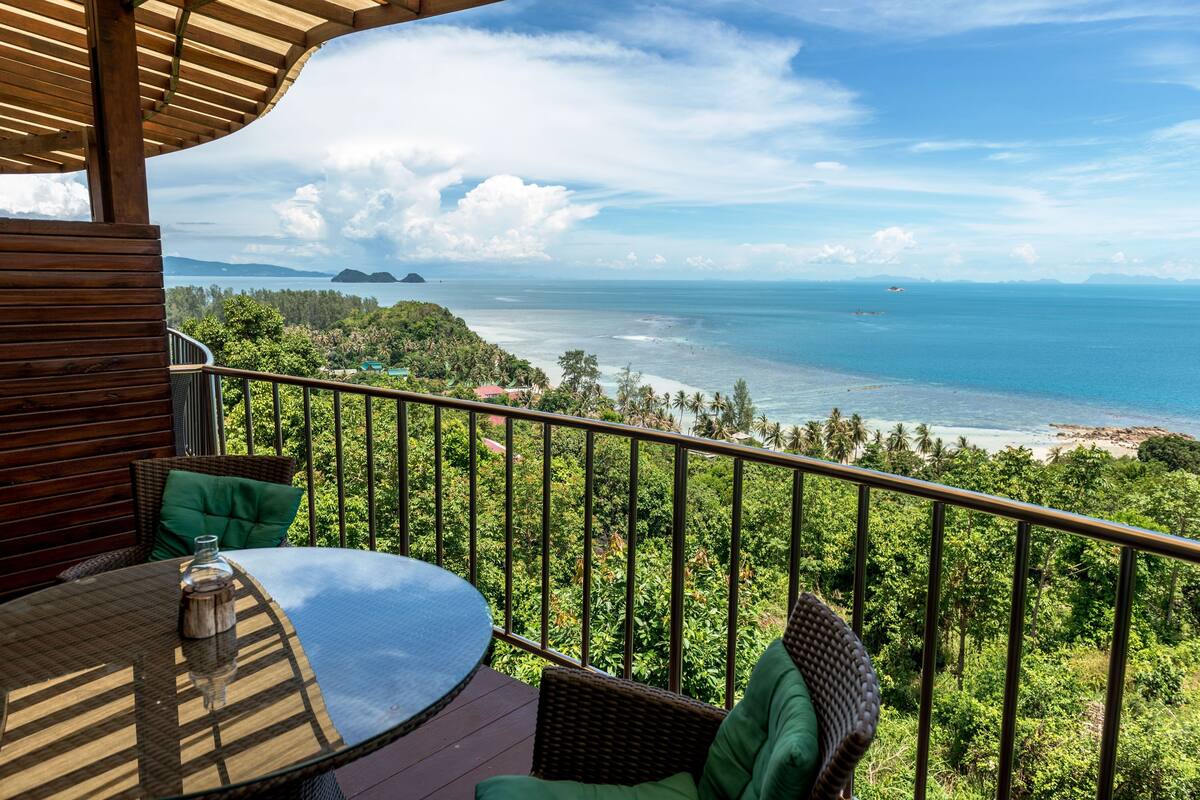 A wooden deck with a glass table and wicker chairs, overlooking a beach and ocean with distant islands.