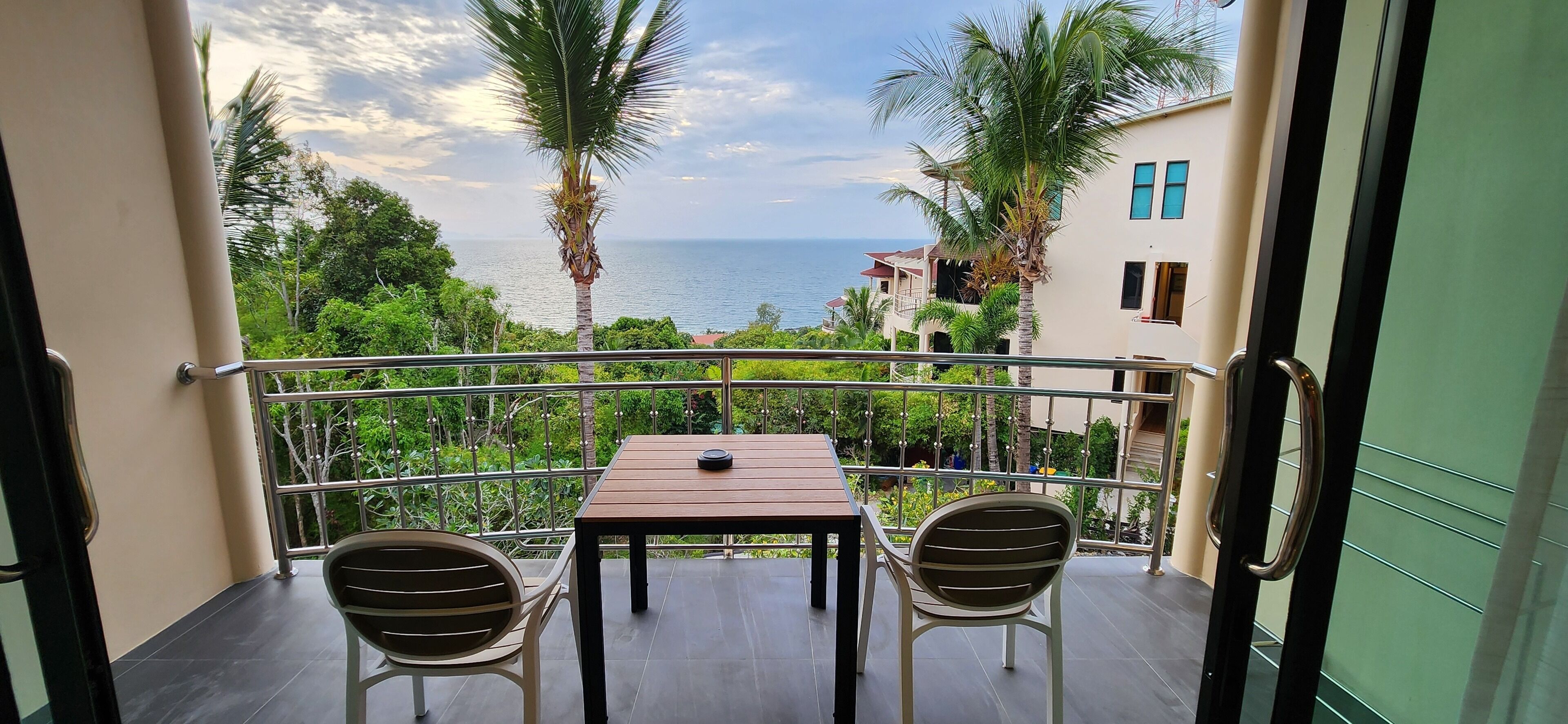 A balcony with two chairs and a table, overlooking a scenic view of the sea and palm trees.