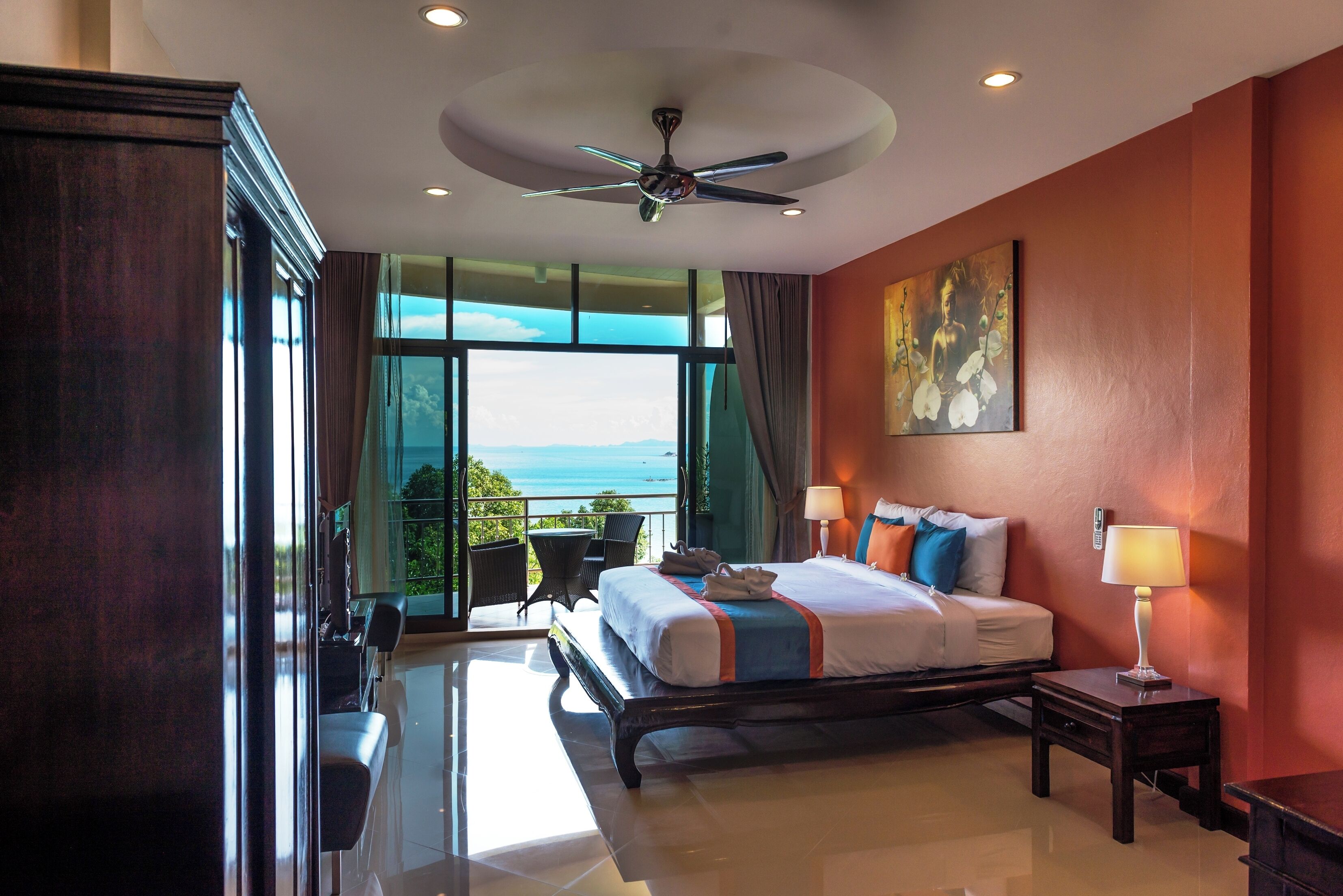 A hotel room with a bed, bedside lamps, a ceiling fan, a large window with a view, and a painting on the wall.