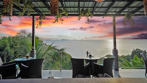 An outdoor seating area with wicker chairs and tables, overlooking a scenic ocean view at sunset.