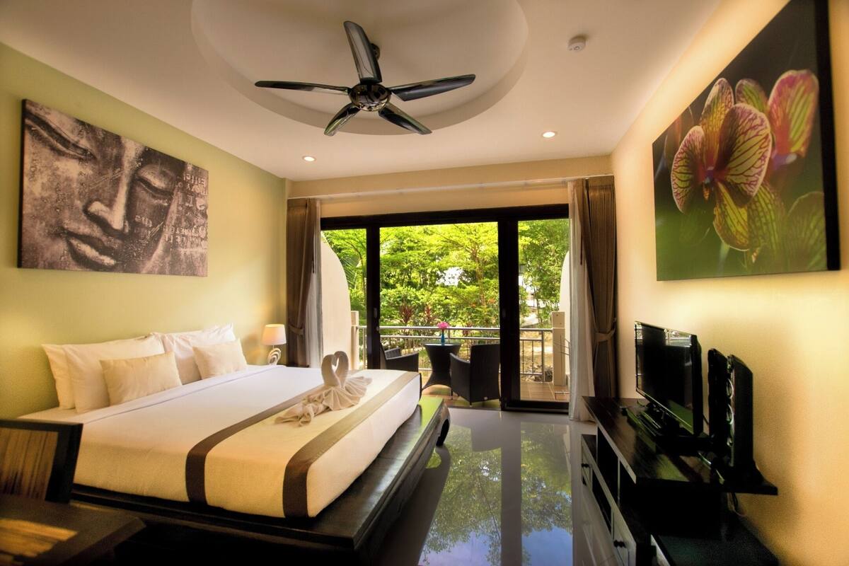 A modern hotel room with a large bed, a ceiling fan, a TV, and a balcony with a view of greenery.