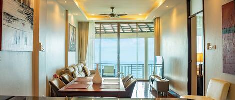 A modern hotel room with a large dining table, comfortable seating, a ceiling fan, and a view of the sea through large windows.