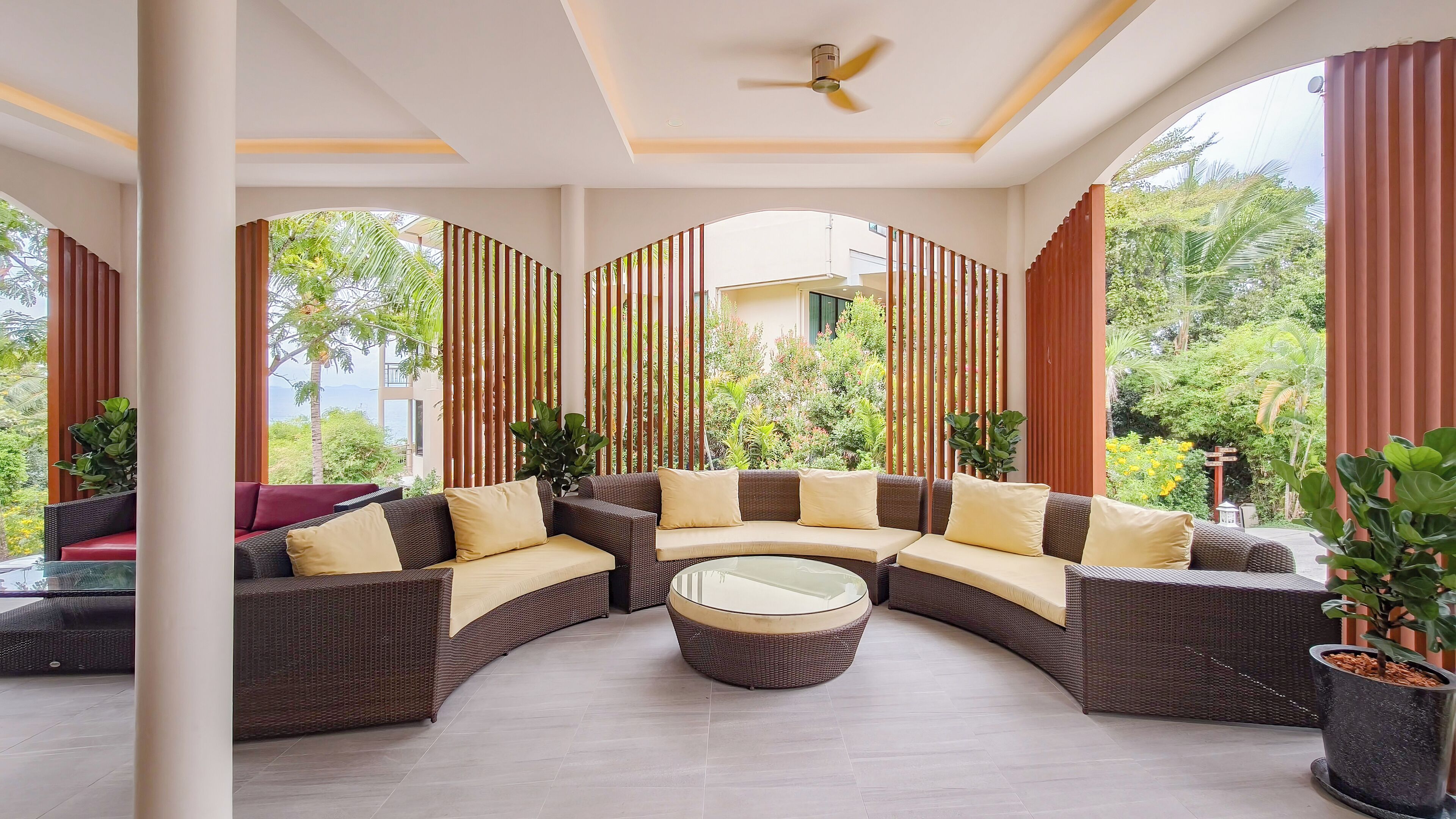 A spacious outdoor seating area with a curved sofa and a central coffee table, surrounded by vertical bamboo panels and potted plants.