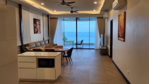A modern hotel room with a dining area, a kitchenette, and a large sliding glass door leading to a balcony with a view of the sea.