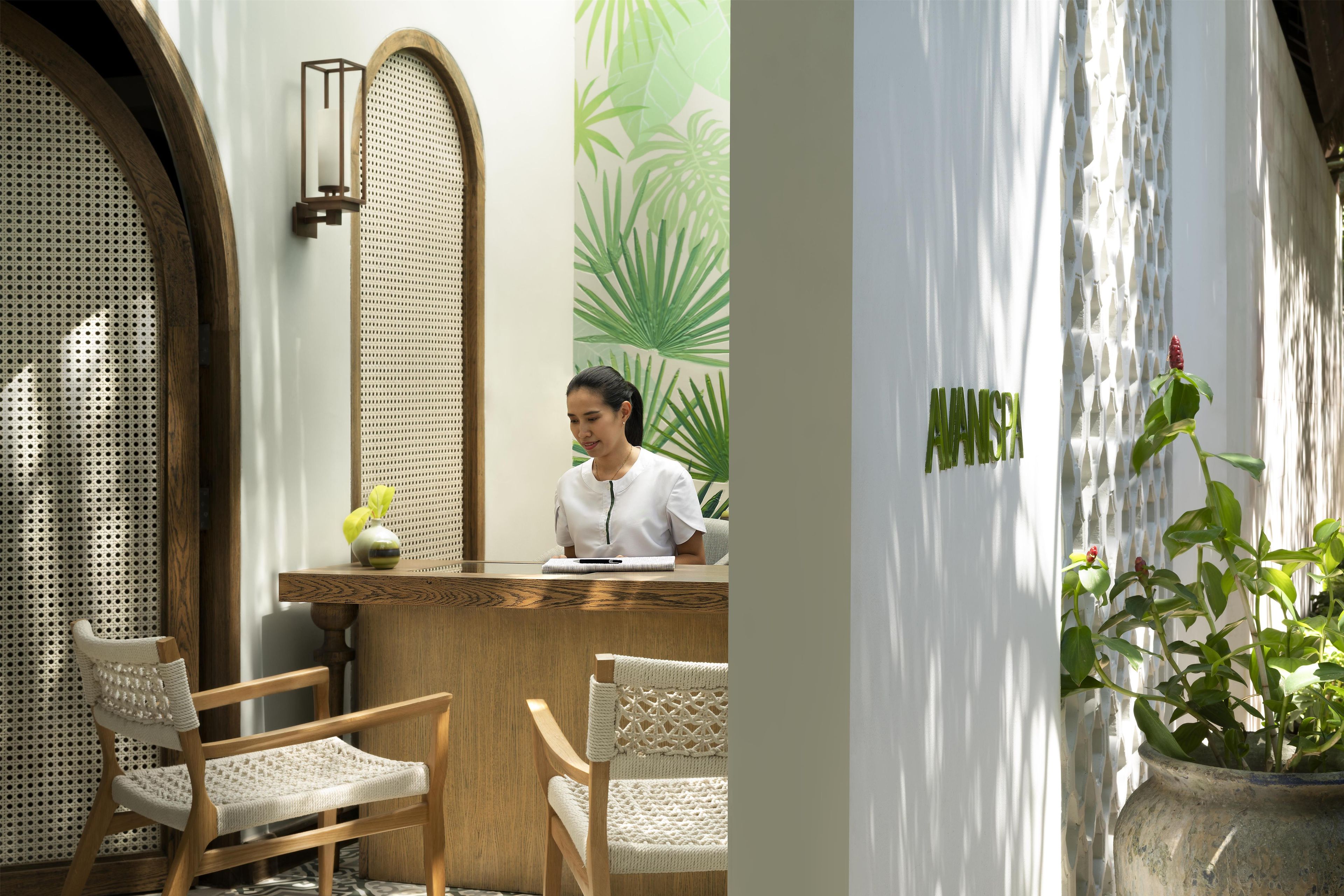 A reception area with a wooden desk, two wicker chairs, and a large green plant in the background.