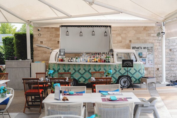 An outdoor seating area with tables and chairs, a food truck with a menu board, and a brick wall in the background. - Avani+ Vacation Club Samui (Koh Samui)