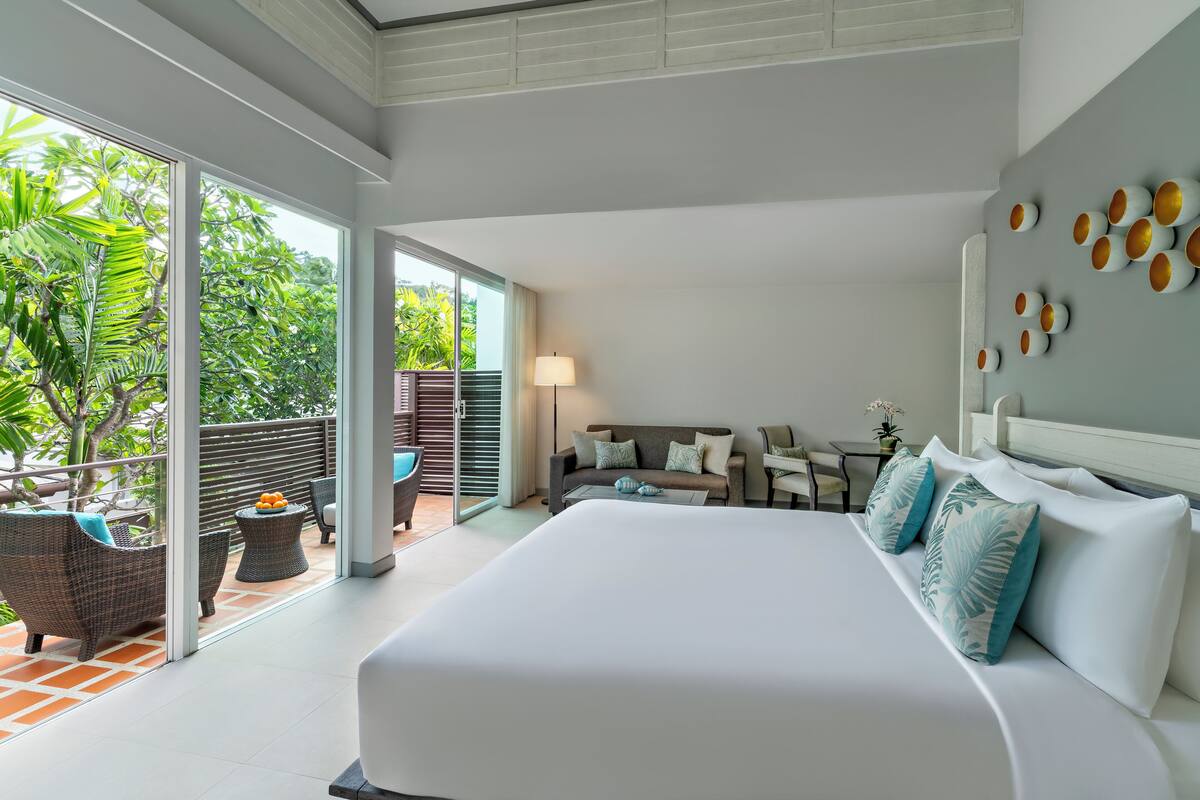 A spacious bedroom with a large bed, a balcony with wicker furniture, and a view of lush greenery.