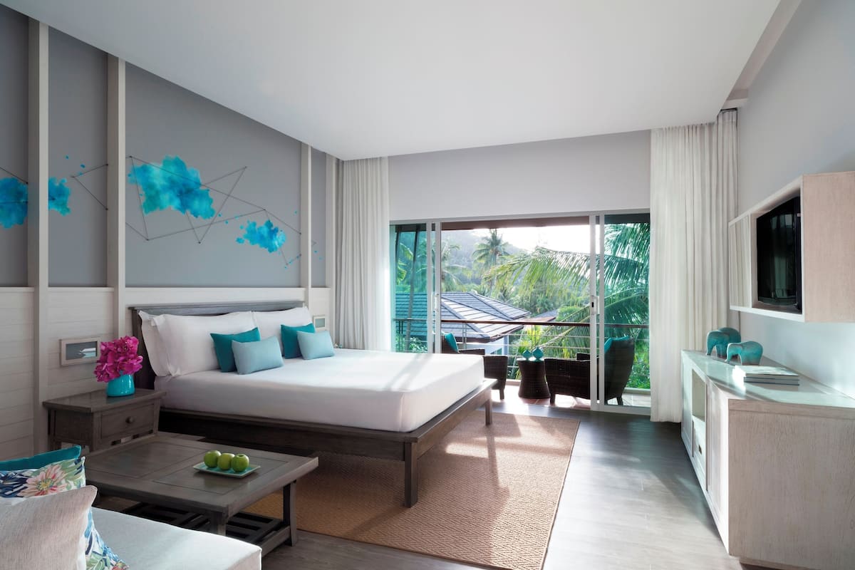 A modern hotel room with a large bed, a TV, a dining table with apples, and a balcony with a view of palm trees.