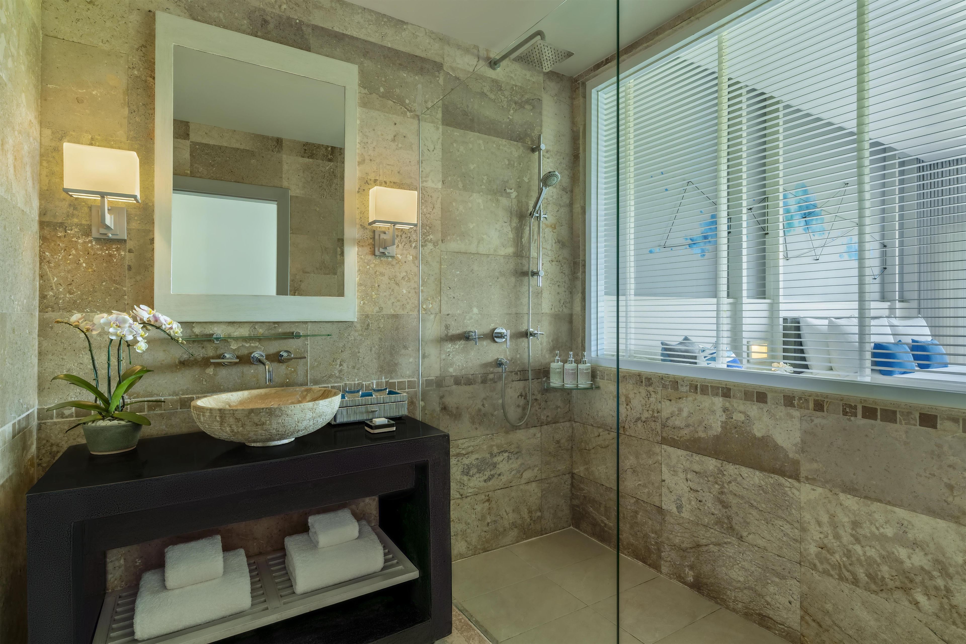 A modern bathroom with a stone wall, a glass shower enclosure, a large mirror, and a wooden vanity with a bowl sink.