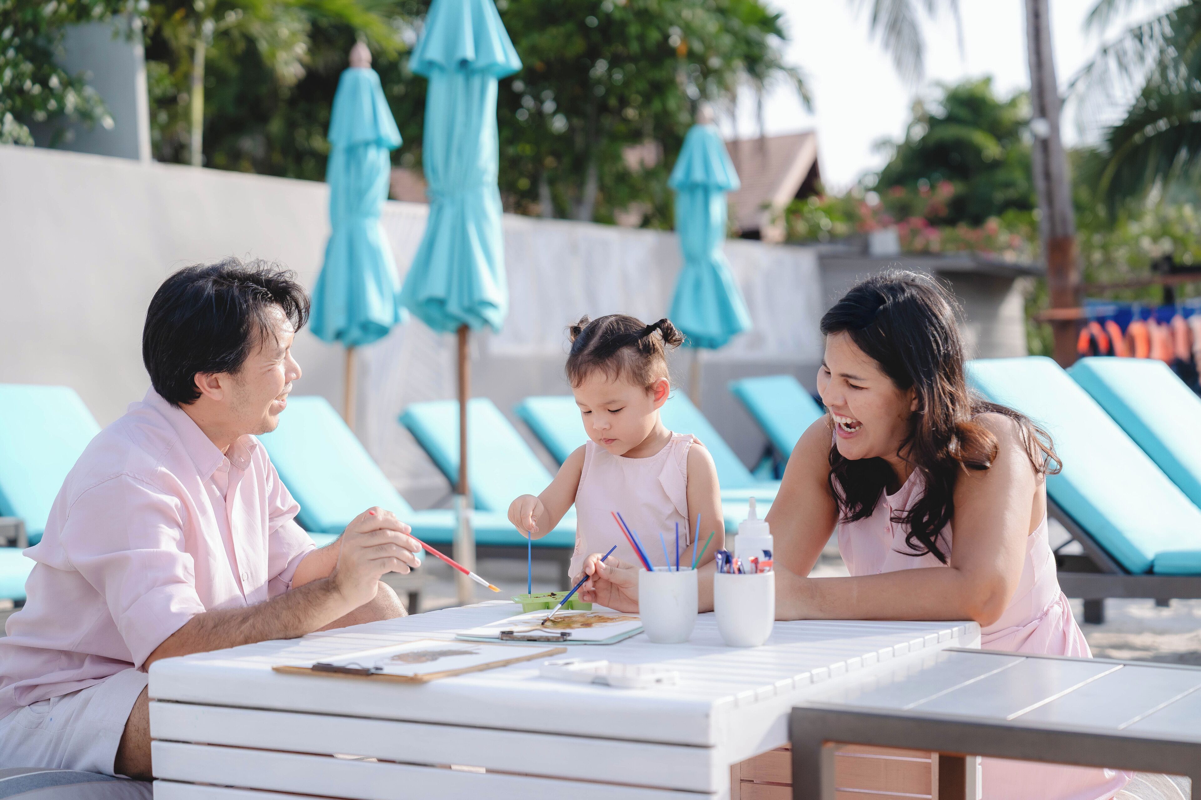 A family of three at a poolside table with a painting activity, surrounded by lounge chairs and umbrellas.