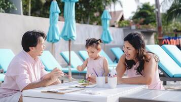 A family of three at a poolside table with a painting activity, surrounded by lounge chairs and umbrellas.