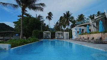A swimming pool with lounge chairs and umbrellas, surrounded by palm trees and a building with a glass facade.
