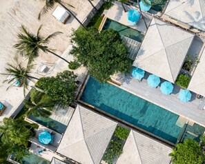 An aerial view of a resort with a central swimming pool, multiple lounge areas with umbrellas, and surrounding palm trees.