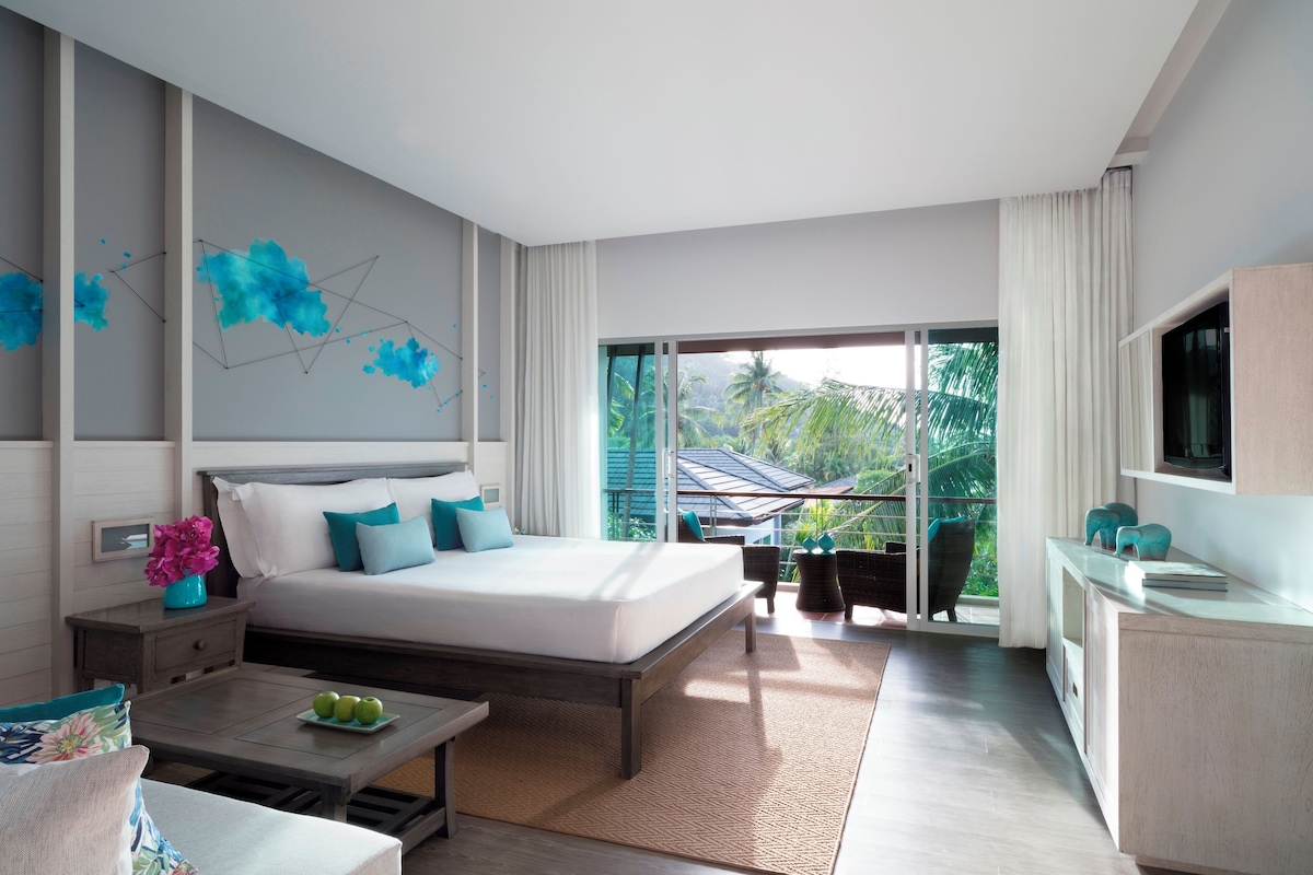 Una habitación de hotel moderna con una cama grande, un televisor, una mesa de comedor con manzanas y un balcón con vistas a palmeras.