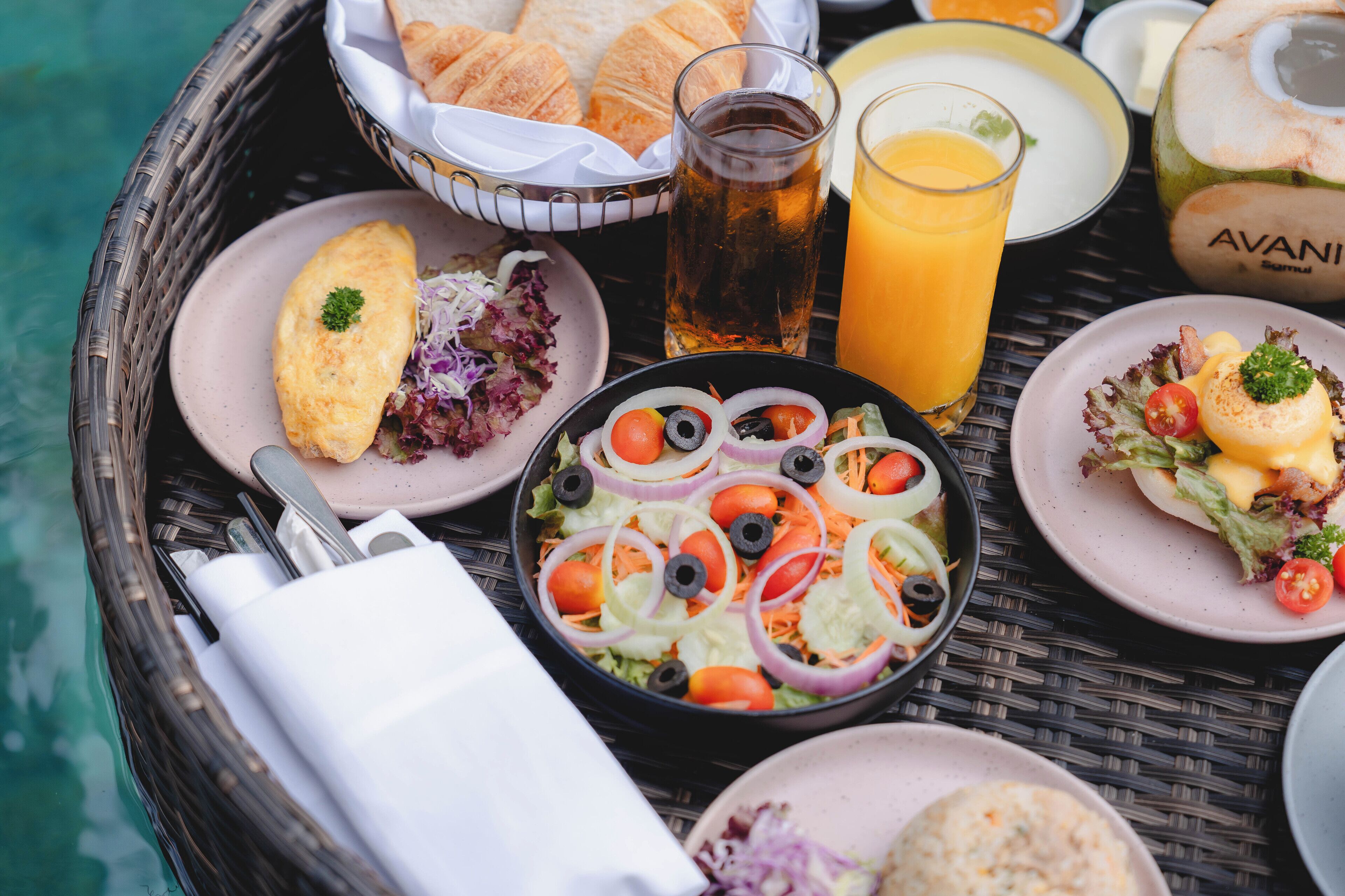 Un plateau en osier contenant des aliments pour le petit-déjeuner, notamment des croissants, une salade, des œufs et du jus de fruits.