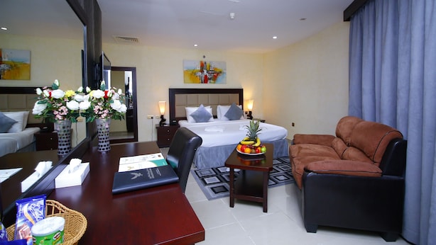 A hotel room with a bed, a sofa, a desk with a laptop, a fruit bowl, and a vase with flowers. - Royal Falcon Hotel (Dubai)