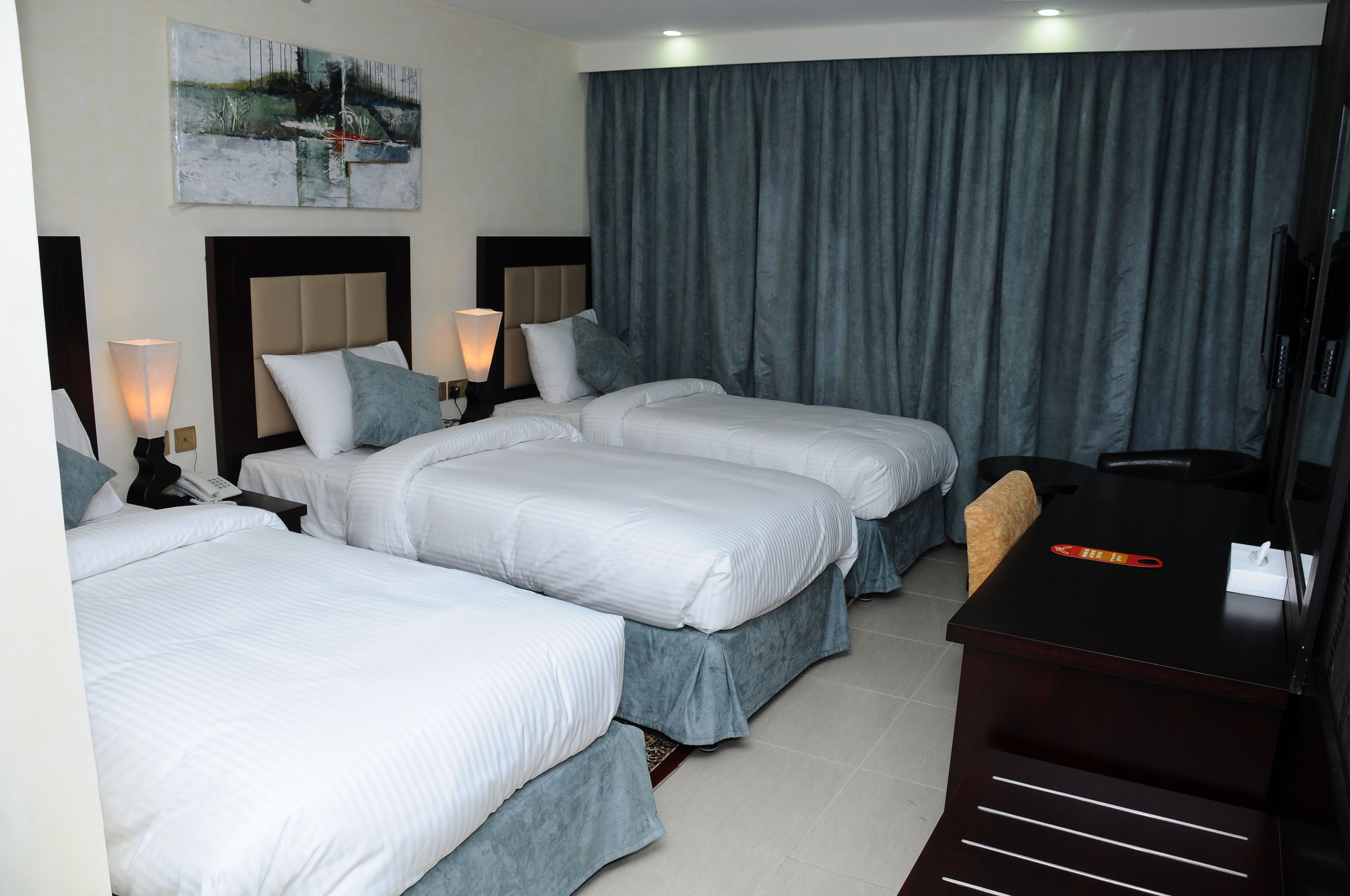 A hotel room with three beds, a desk, a TV, and a painting on the wall.