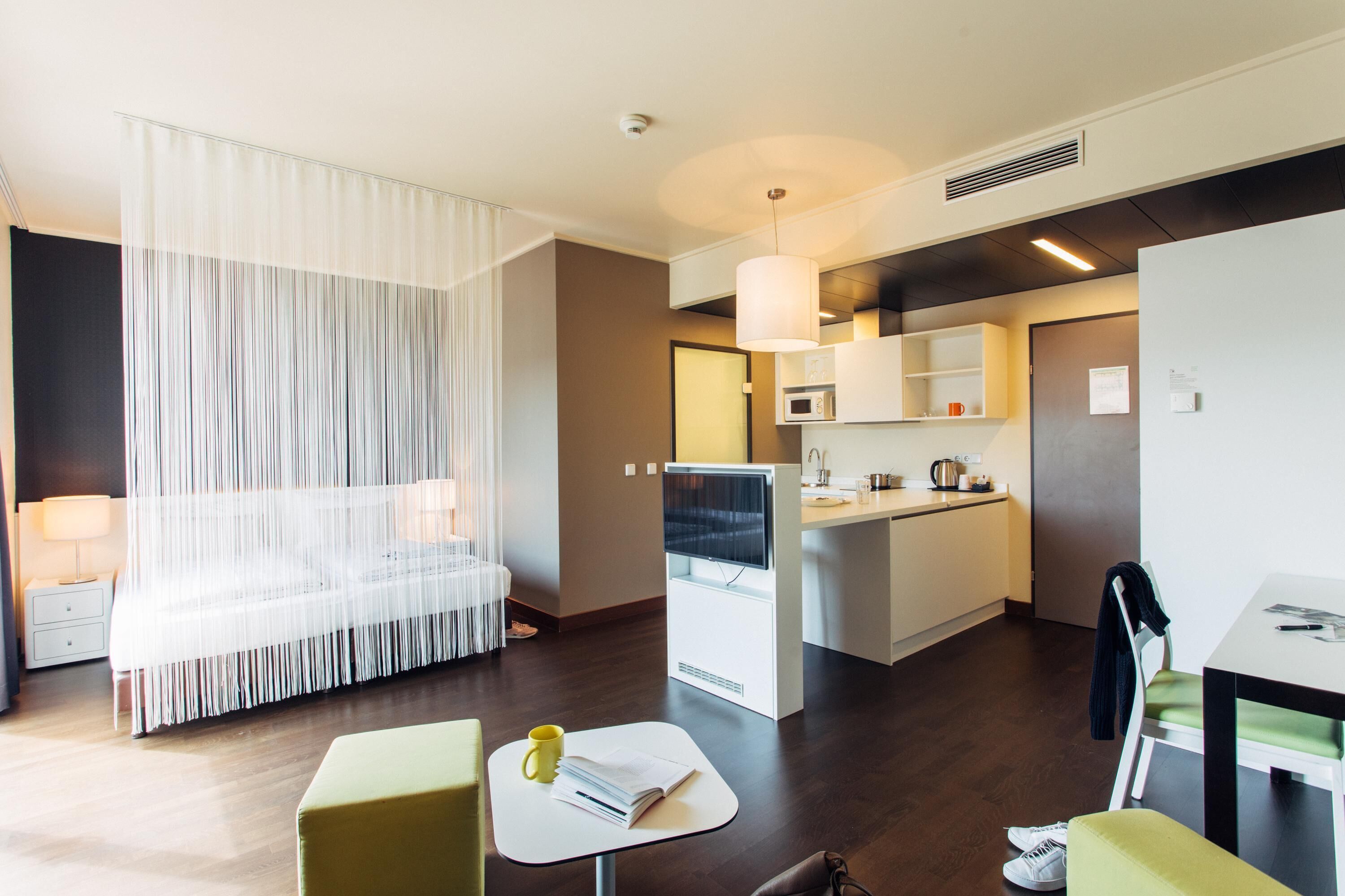 A modern hotel room with a bed, a small table with a book and a mug, a kitchenette, and a dining area.