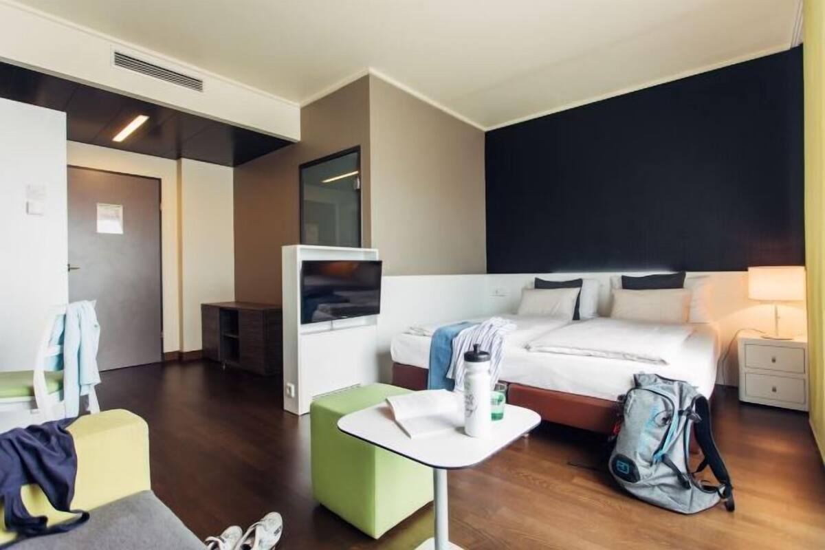 A hotel room with a bed, a small table, a chair, a TV, a desk, and a wardrobe.