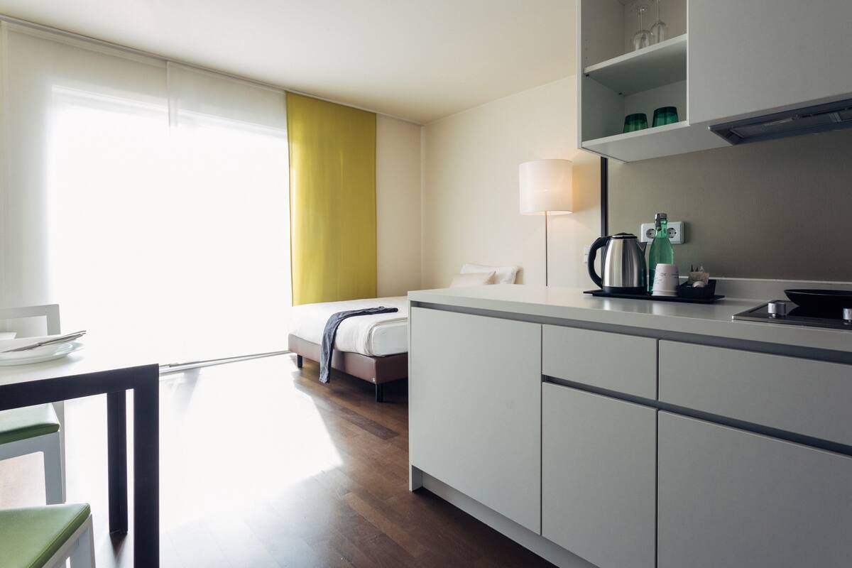 A modern hotel room with a kitchenette, a bed with a green curtain, a dining table with chairs, and a view of the outdoors.