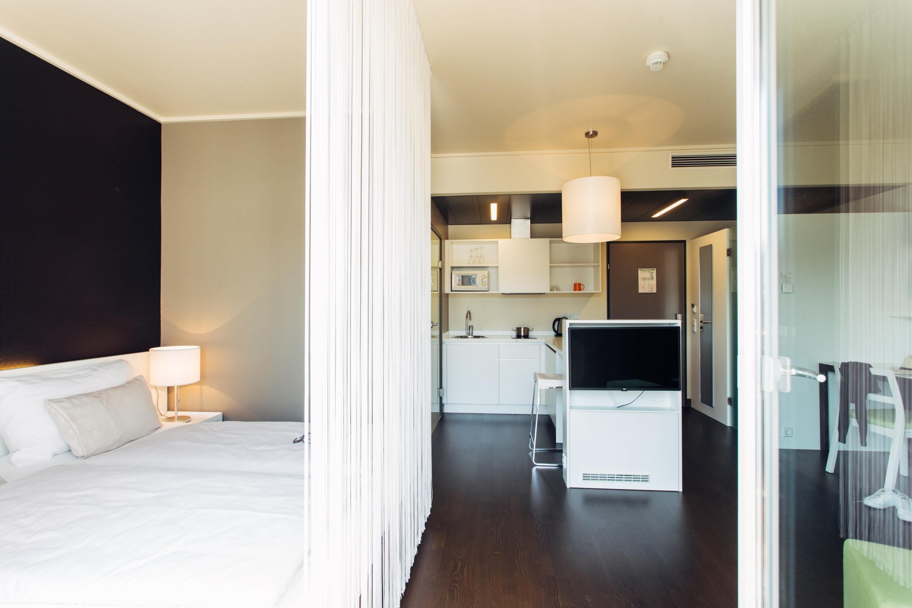 A hotel room with a bed, a desk, a chair, a TV, and a kitchenette.