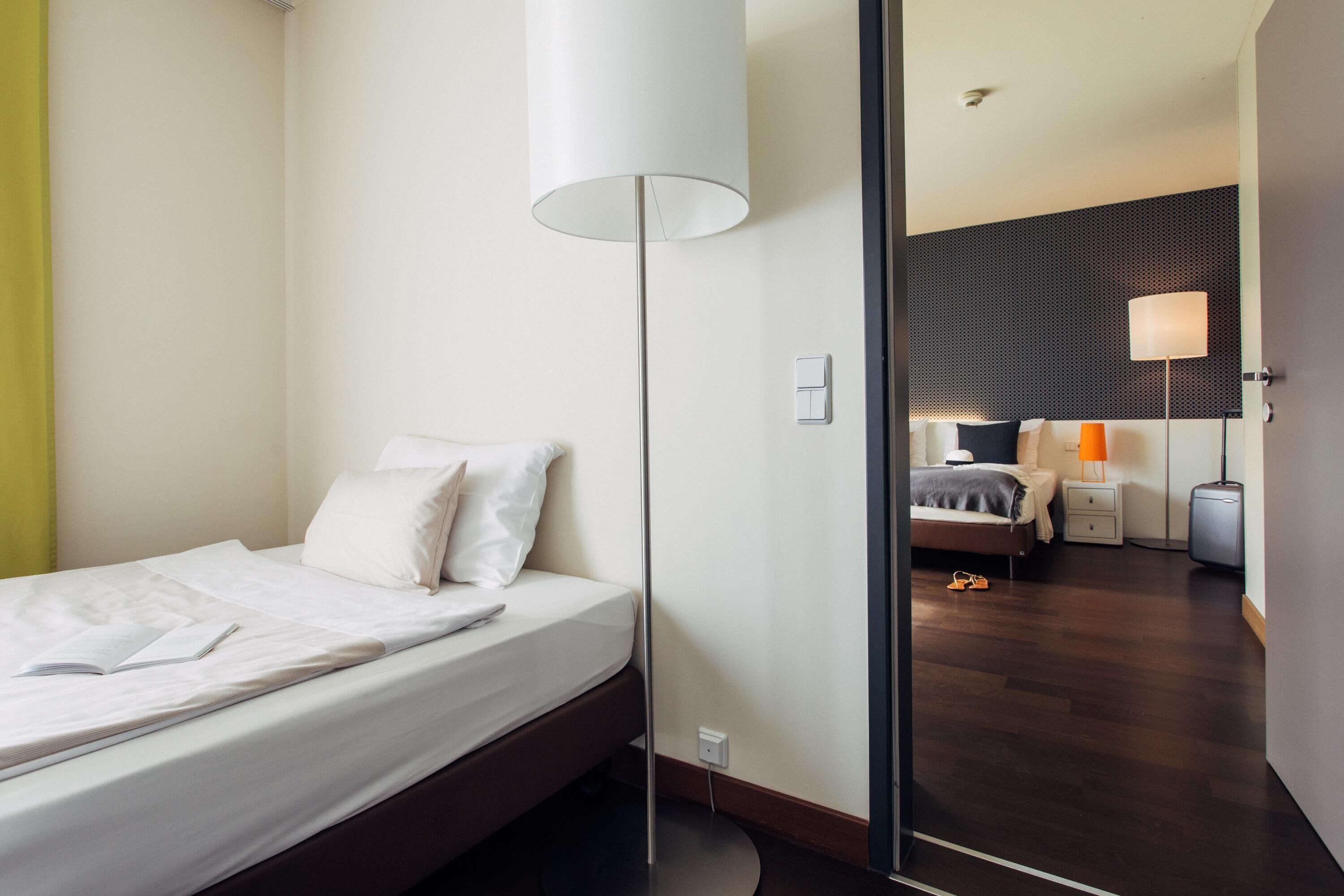A modern hotel room with a neatly made bed, a standing lamp, and a view into an adjacent room with a similar setup.