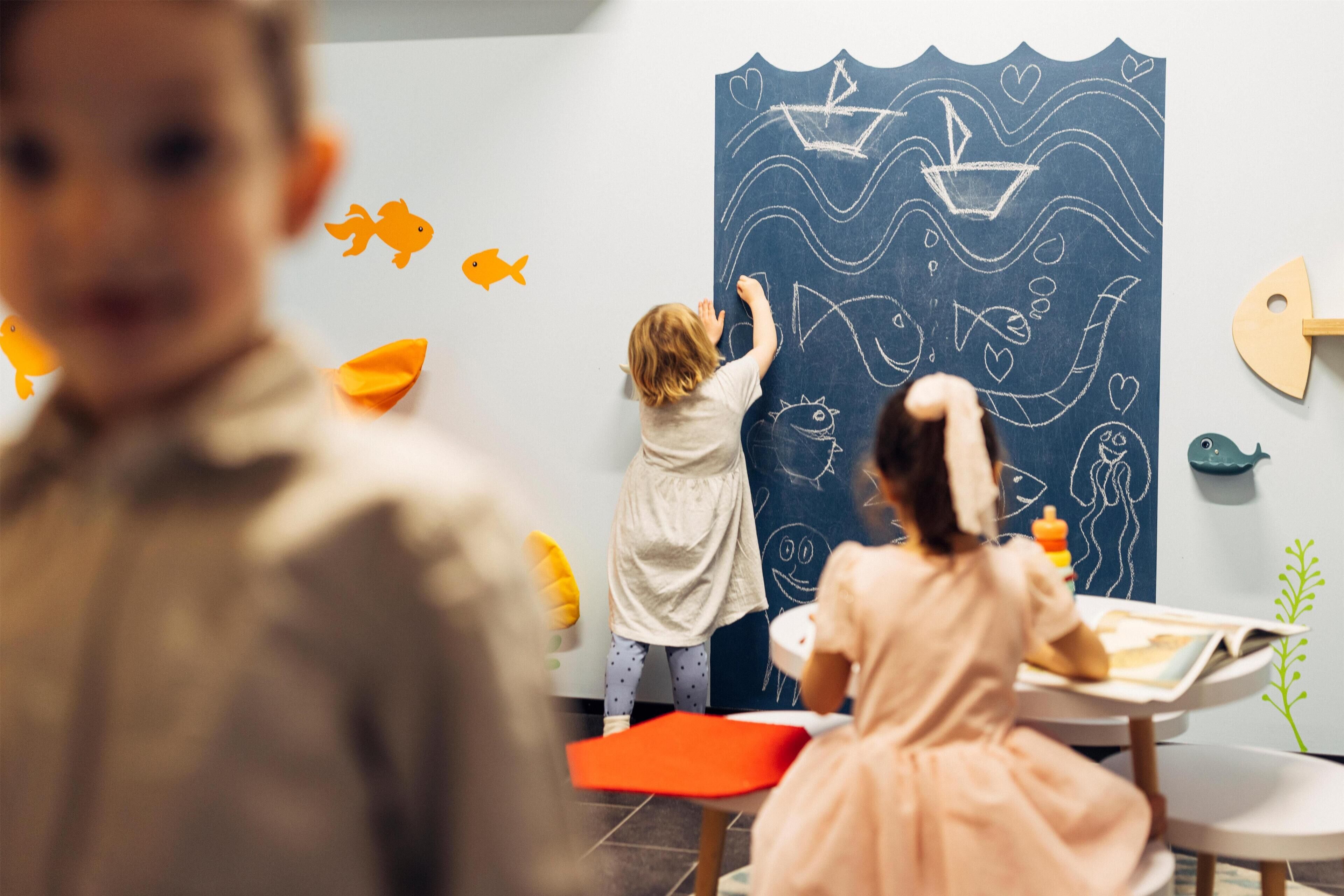 Children drawing on a chalkboard, with a wooden fish decoration and a plant on the wall.