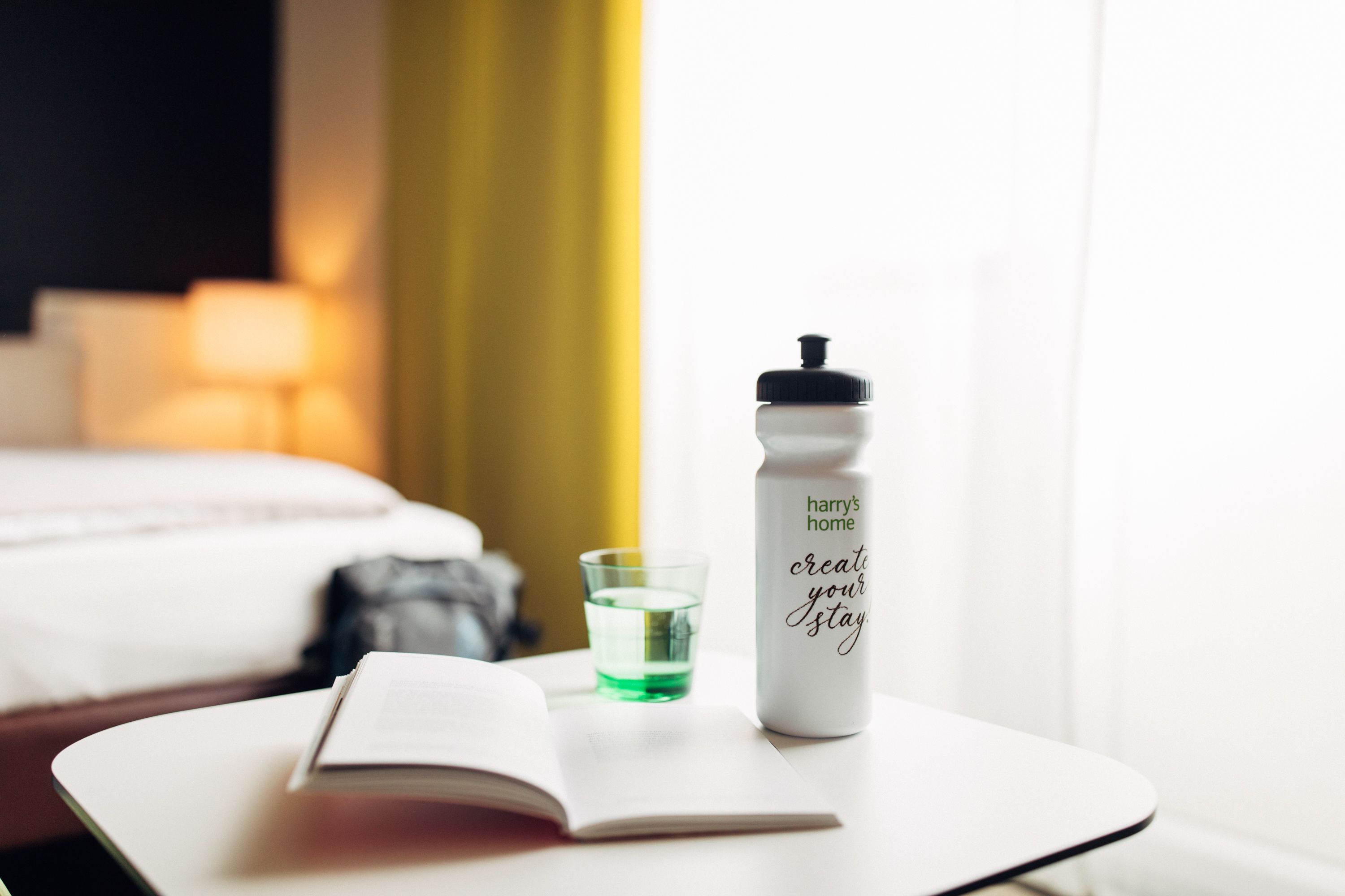 A hotel room with a bed, a bedside table with a book and a water bottle, and a glass of water.