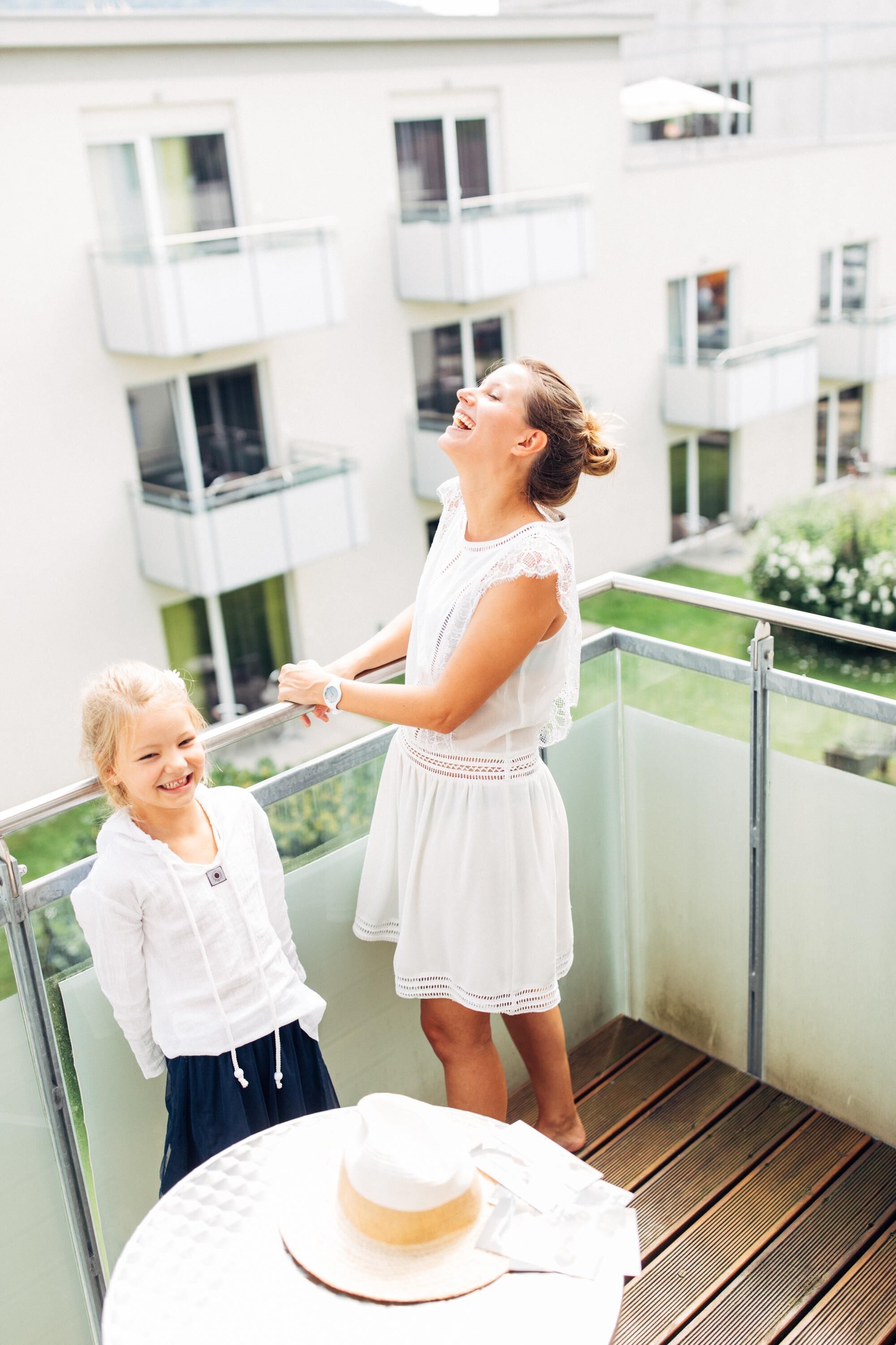 Un balcon avec une balustrade, une table sur laquelle se trouve un chapeau, et un bâtiment avec des balcons en arrière-plan.