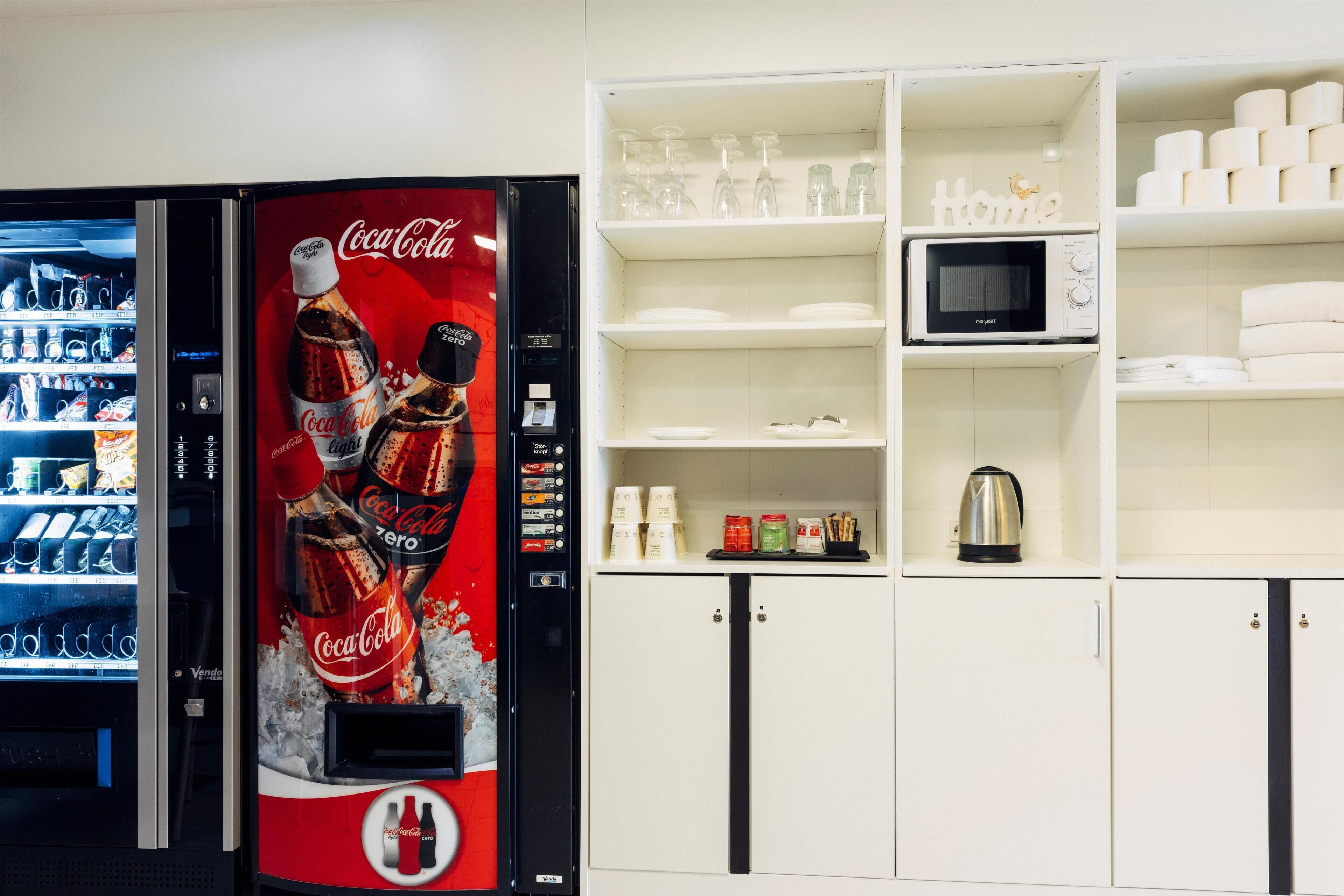 Máquina de vendas com marca Coca-Cola, um micro-ondas e prateleiras com vários itens, incluindo pratos e copos.