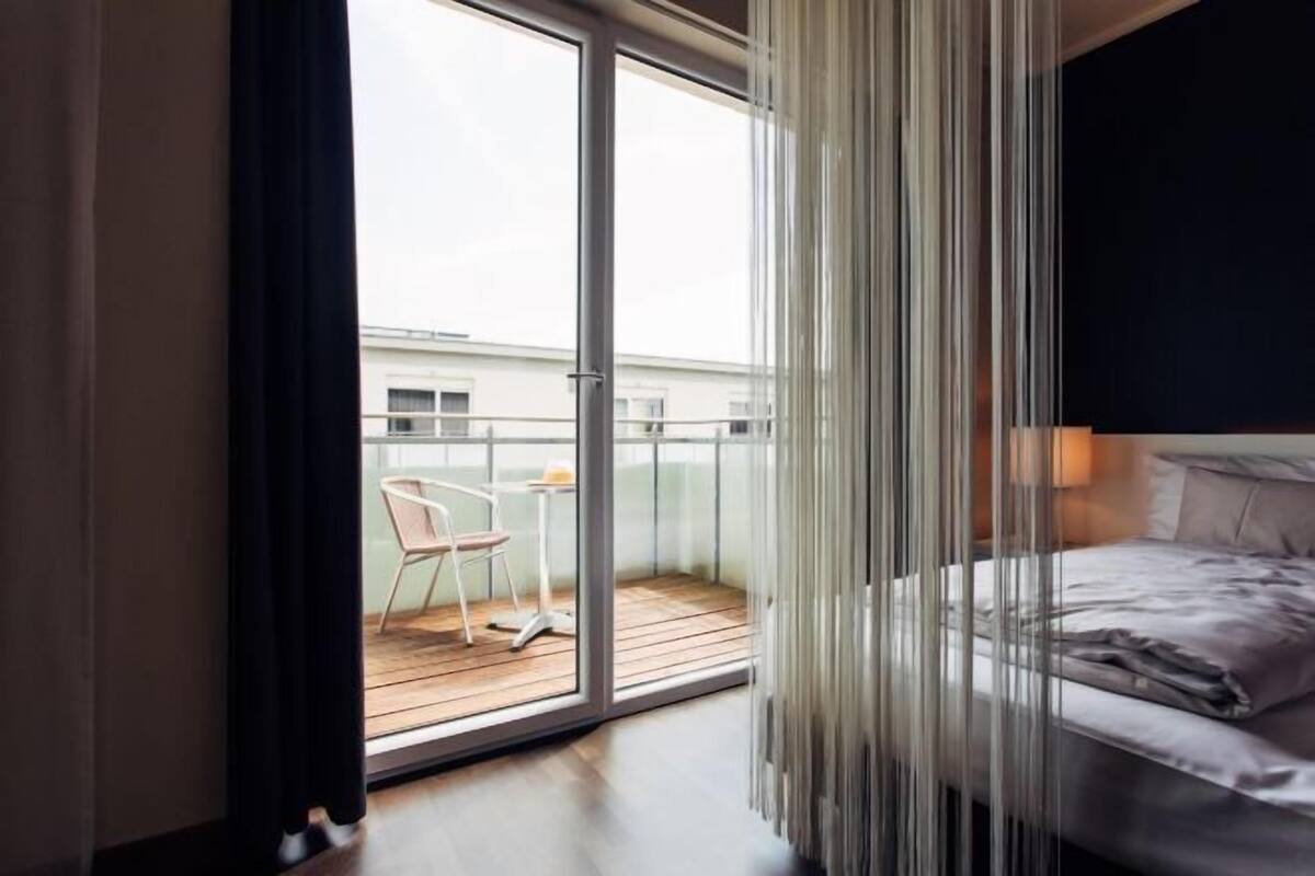A bedroom with a bed, a window, and a view of a balcony with a chair and table.