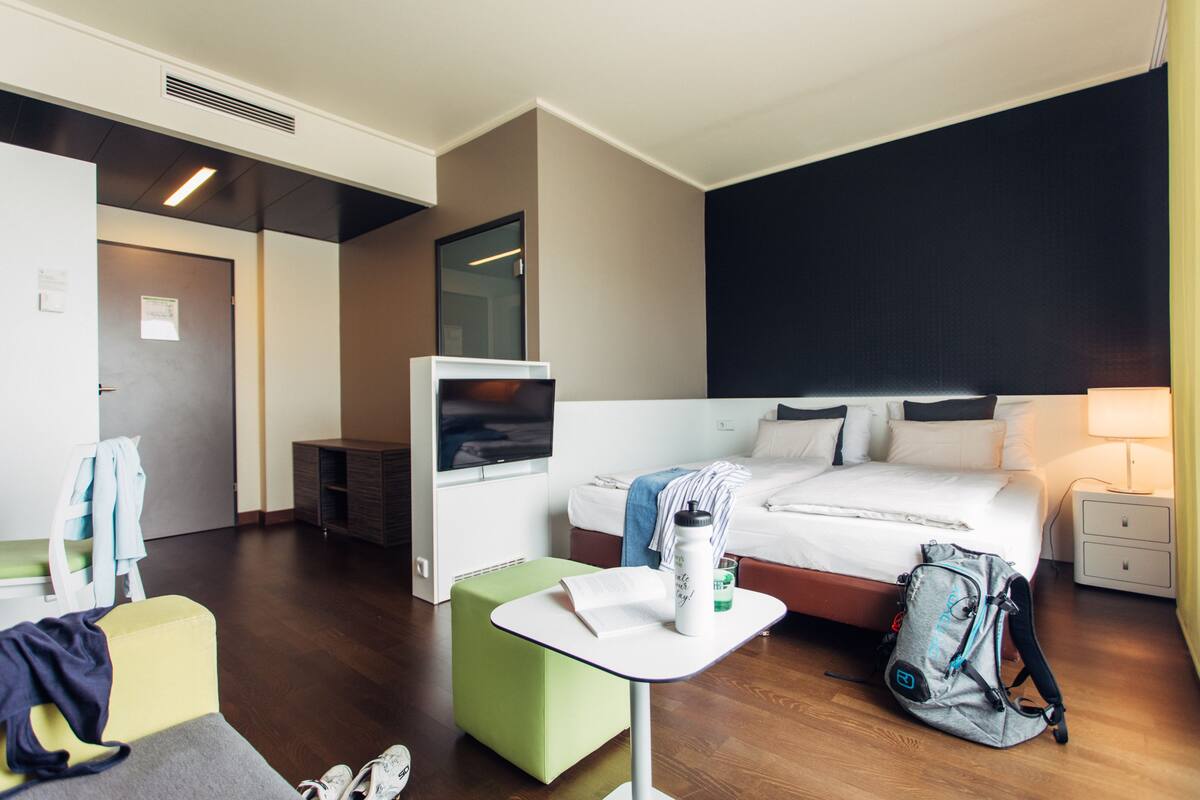 A hotel room with a bed, a TV, a small table, a green ottoman, a backpack, and a nightstand with a lamp.