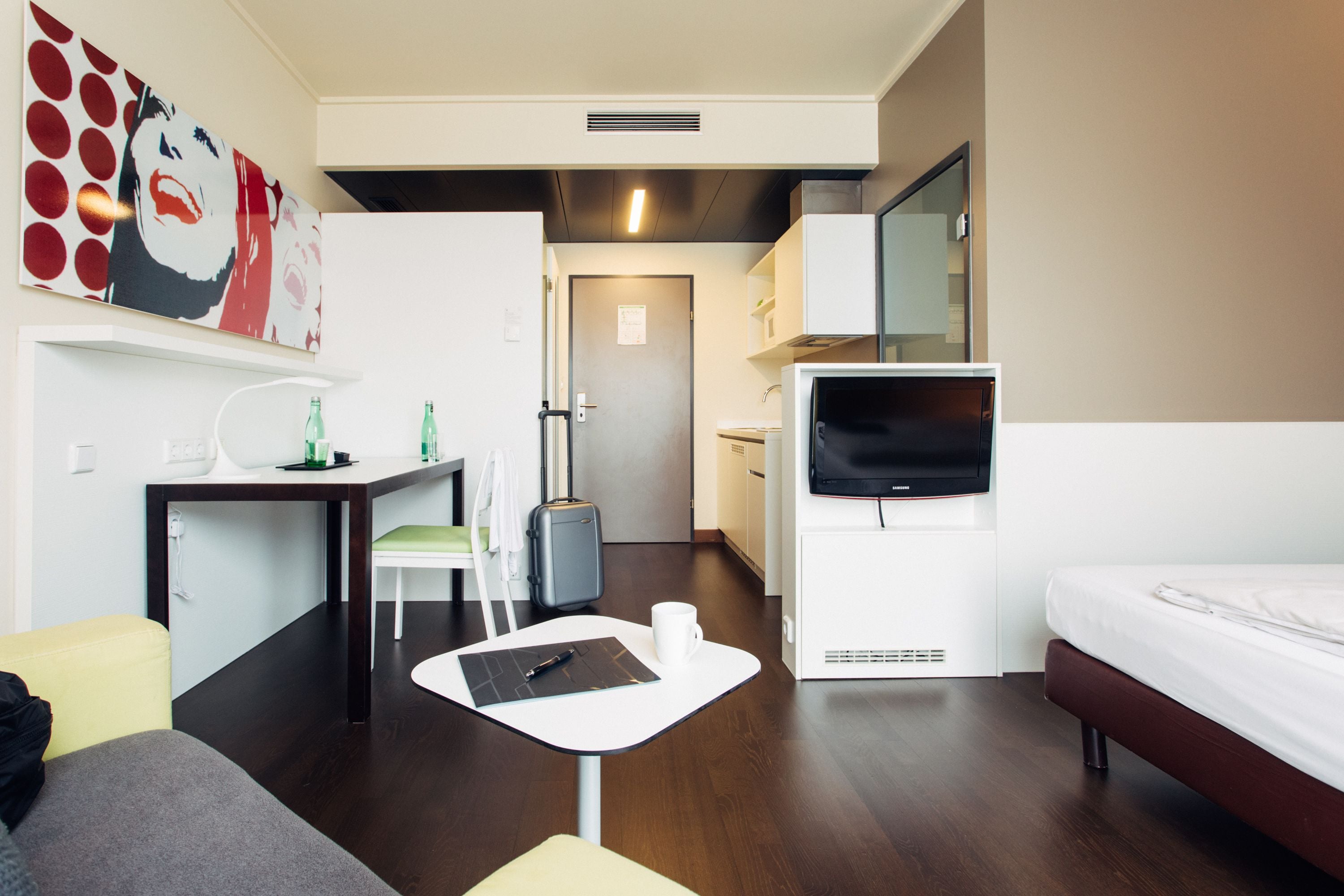A compact hotel room with a bed, a desk with a chair, a small table, a TV, and a bathroom visible through an open door.