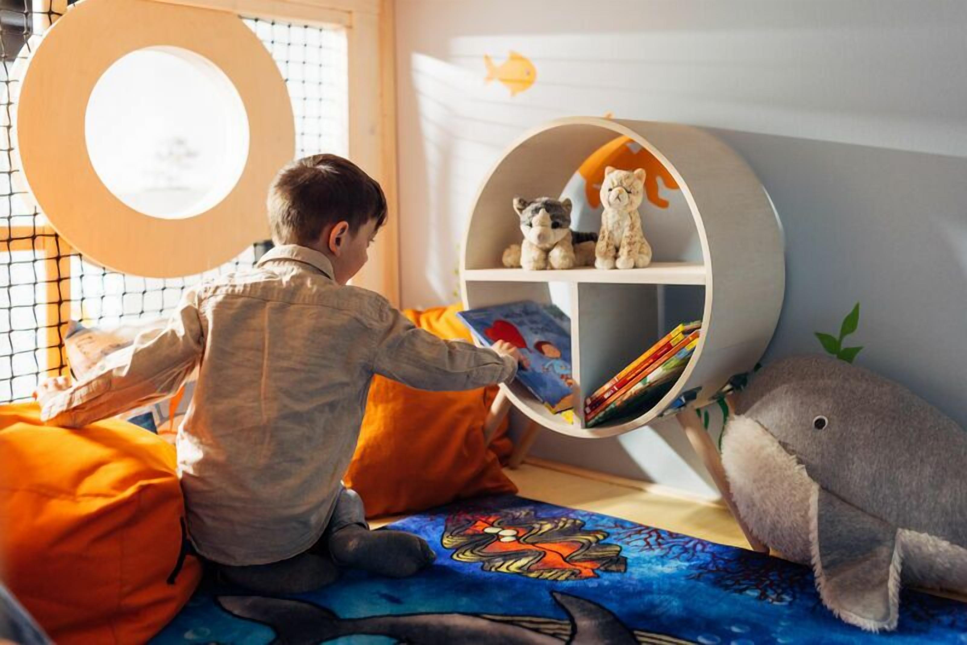 A child reading a book in a room with a bookshelf, a window with a circular frame, and a plush shark toy.