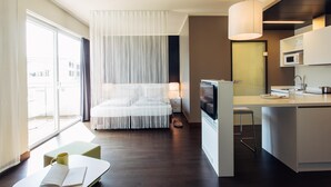 A modern hotel room with a large bed, a TV, a small kitchen area, and a view of the city. - harry’s home Linz-Urfahr hotel & apartments (Linz)