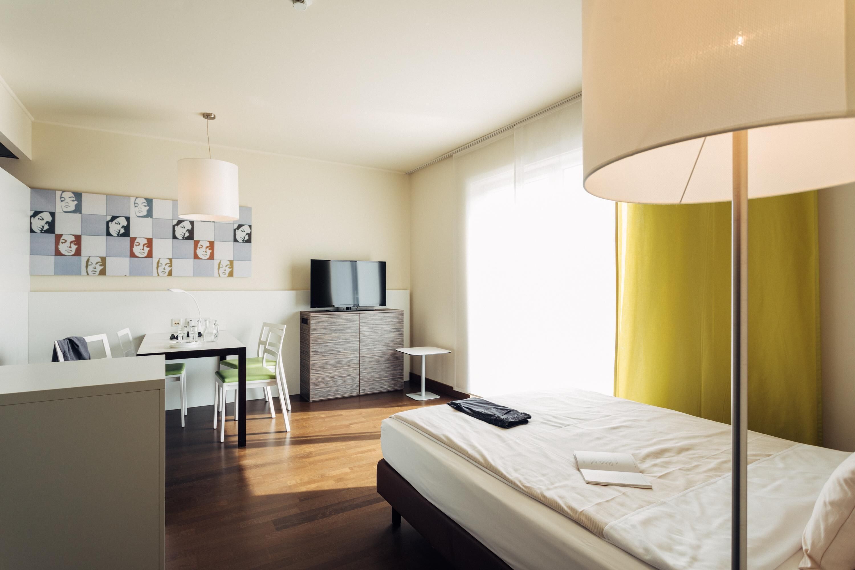 A hotel room with a bed, a desk with chairs, a television, and a wall with a patterned artwork.