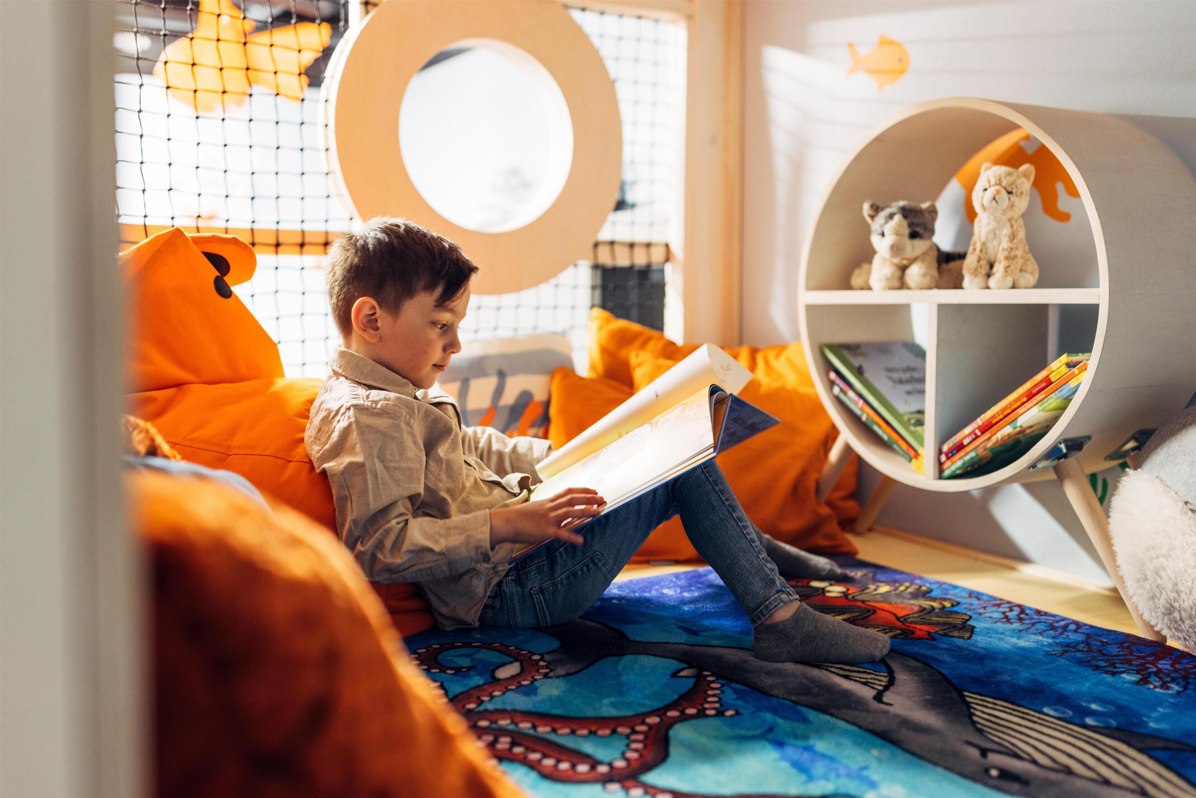 A child reading a book in a cozy room with a bookshelf, a window with a circular decoration, and a colorful octopus-themed blanket.
