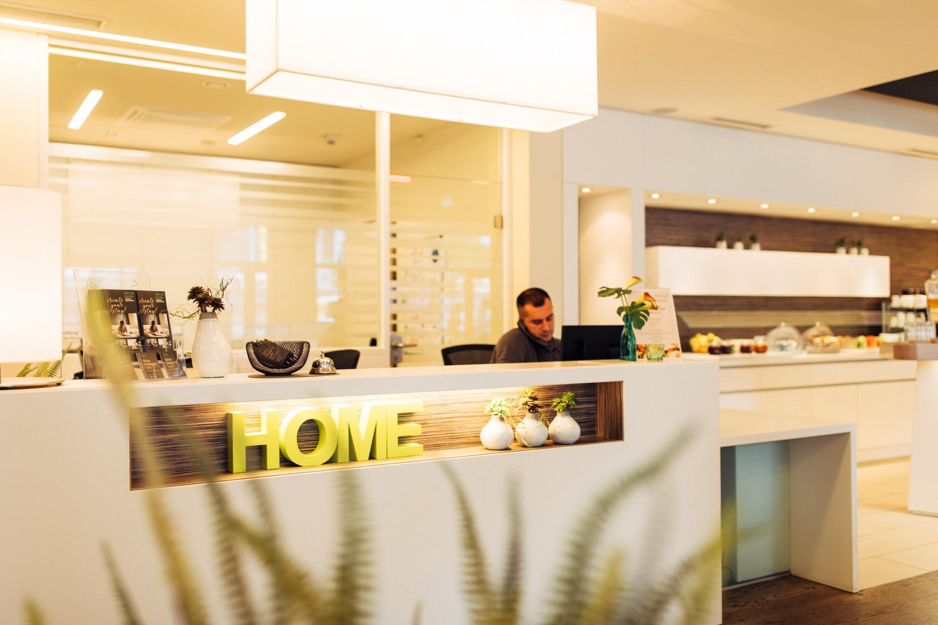 Ein Empfangstresen mit einem 'HOME'-Schild, ein Mann arbeitet an einem Computer, und ein modernes Interieur mit einer Küchenzeile im Hintergrund.